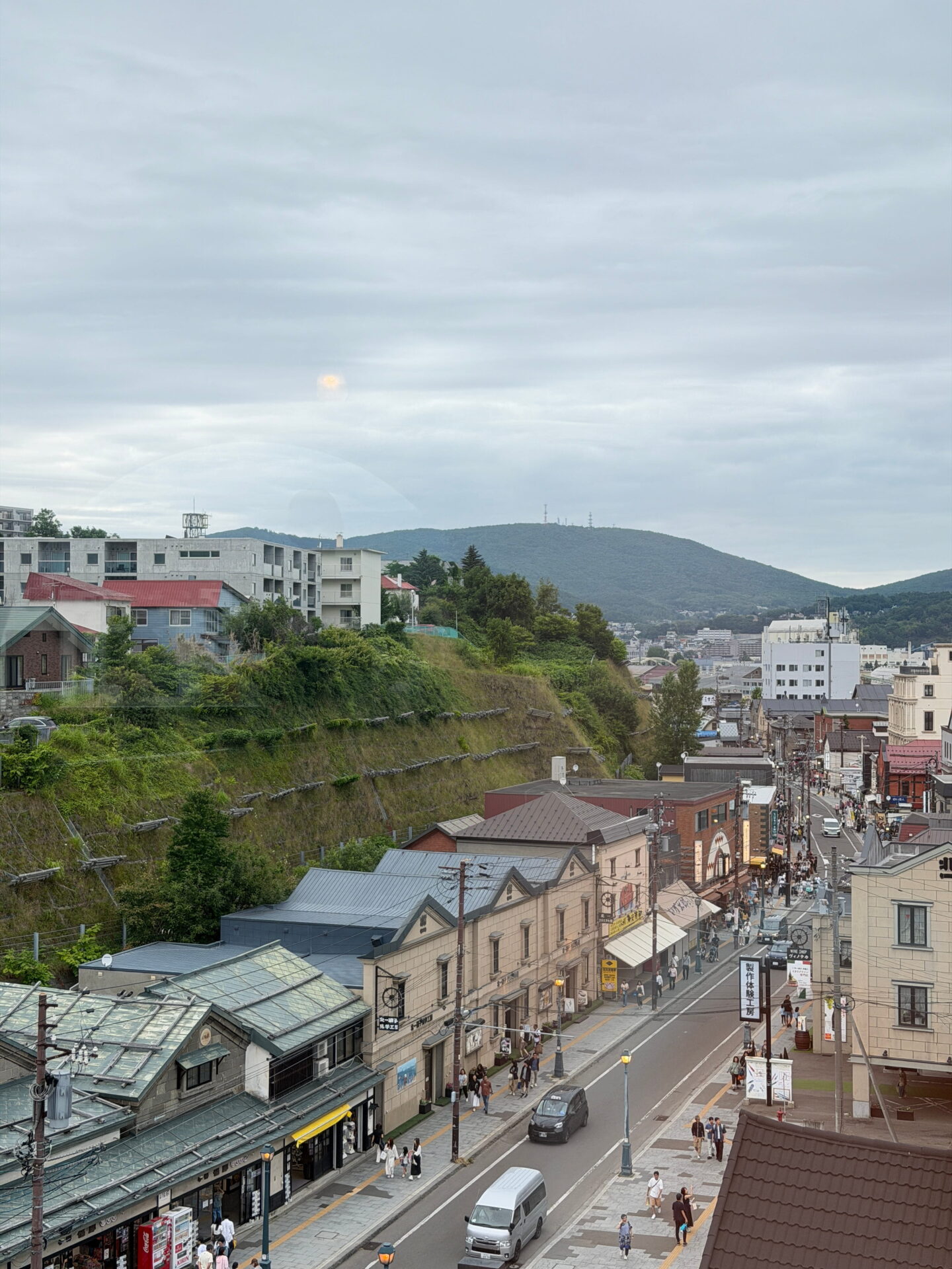 An elevated view over Otaru showing a street lined with historic buildings, shops and pedestrians. A green hillside rises beside the town with forested mountains stretching across the background beneath a cloudy sky.