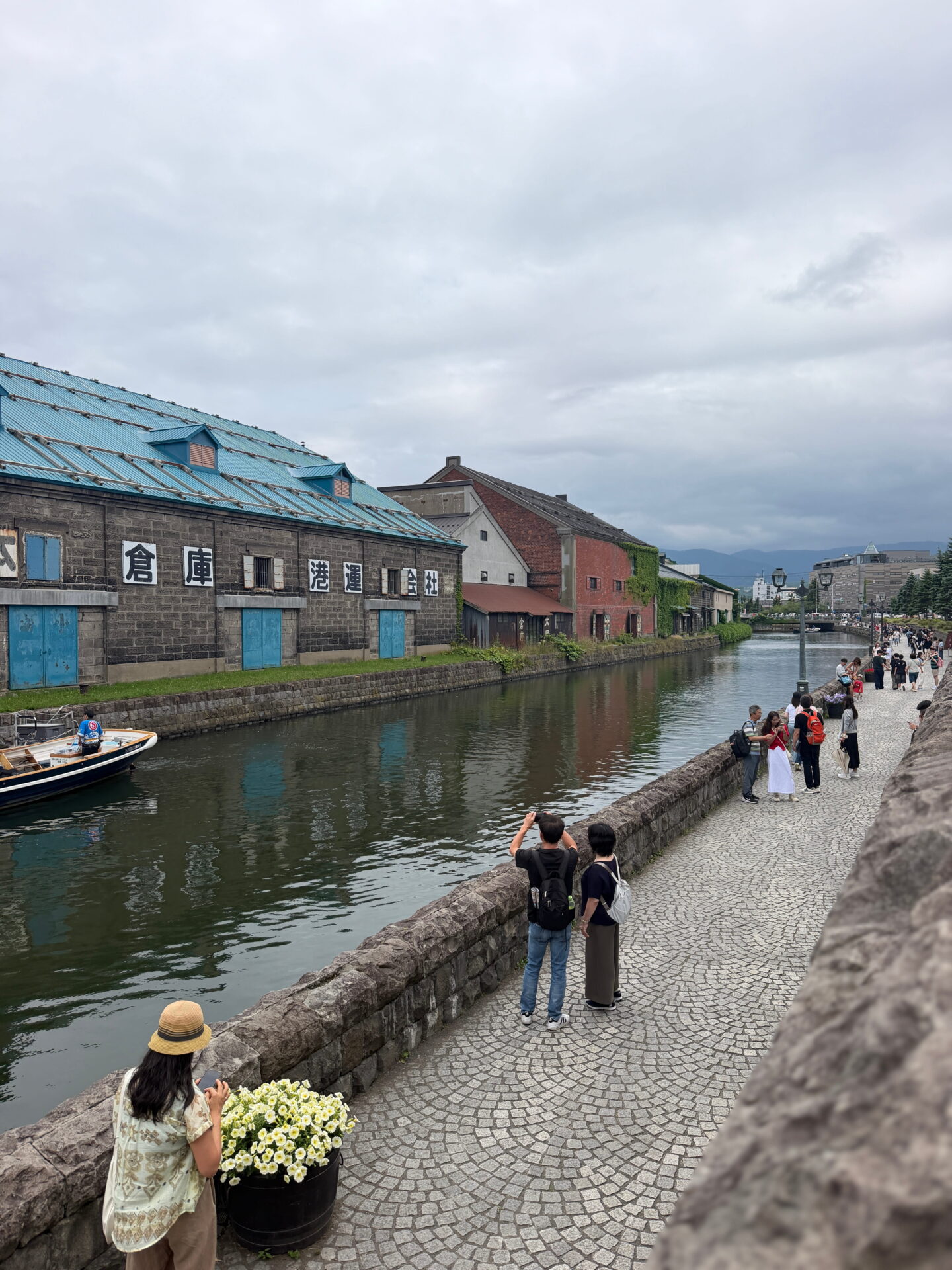 Historic stone warehouses with blue roofs and red brick buildings line Otaru Canal beneath a cloudy sky. Visitors stroll along the cobbled canal path and take photographs beside the water.