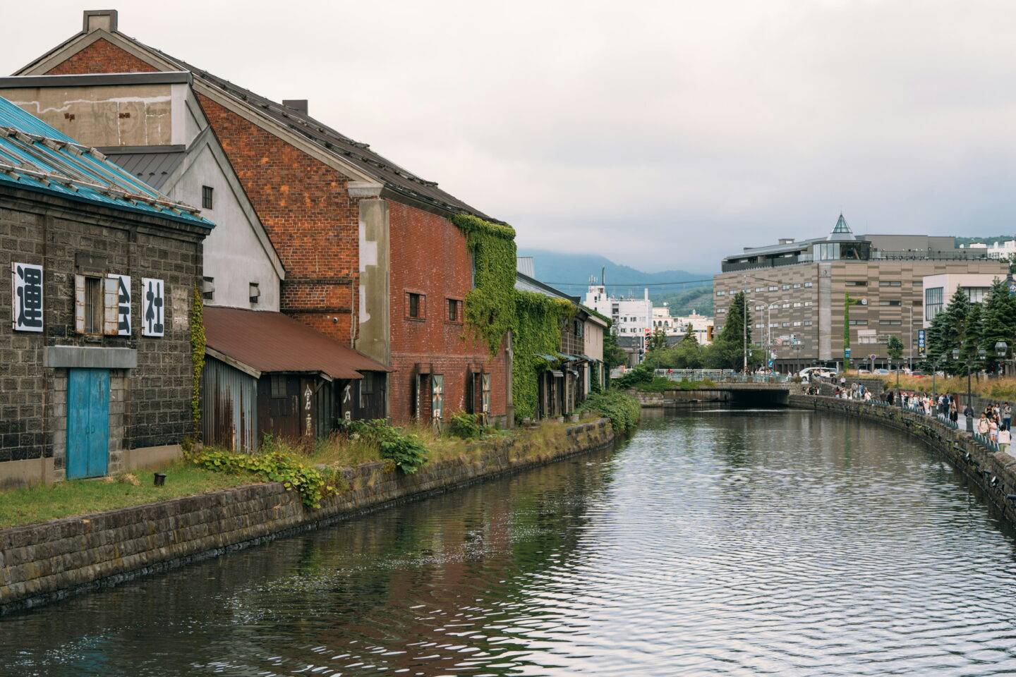Historic stone and red brick warehouses line Otaru Canal in Hokkaido, with bright blue doors and green ivy covering parts of the buildings. The canal curves towards a small bridge while tourists stroll along the waterside promenade beneath an overcast sky.