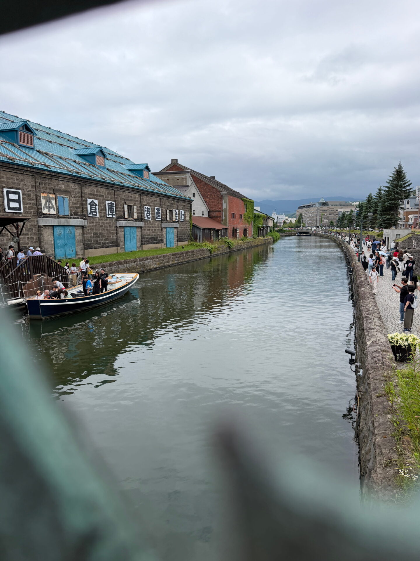 A sightseeing boat carrying passengers travels along Otaru Canal past historic stone warehouses with blue roofs and red brick buildings. Tourists gather along the waterside promenade beneath an overcast sky.