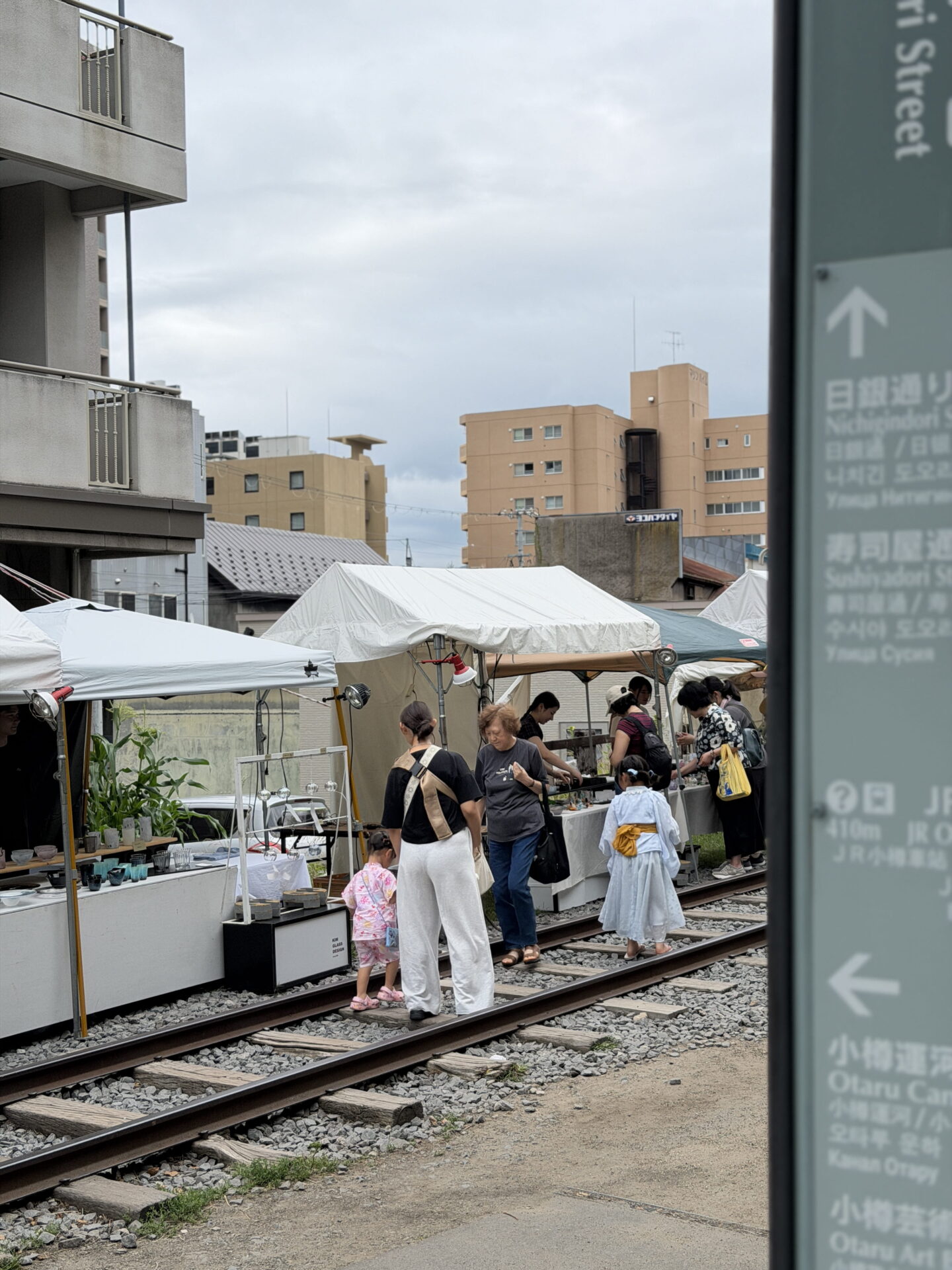 Visitors browse an outdoor glass market set up along disused railway tracks in Otaru. White market tents line the tracks with stalls selling handmade pottery and other small items while families and shoppers walk between them.