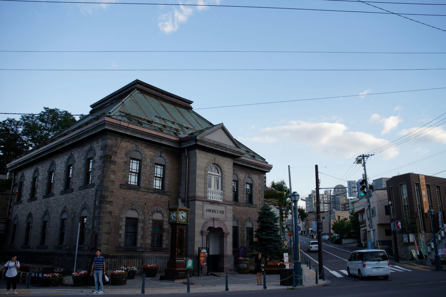 The historic Otaru Music Box Museum in Hokkaido is a two storey brick building with arched windows and a green metal roof. A vintage steam clock stands outside the entrance while pedestrians walk past the building beneath a clear blue sky.