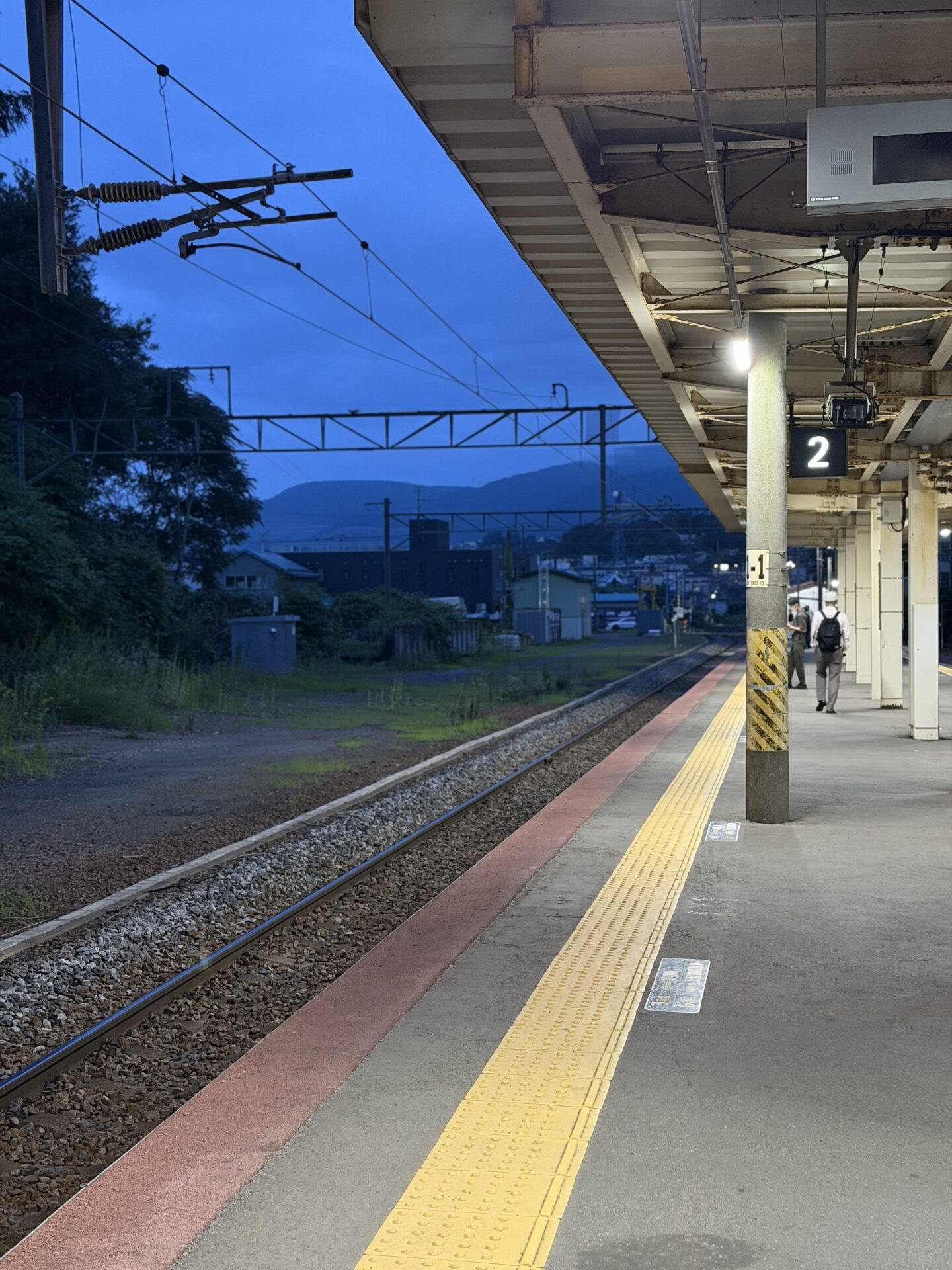 A quiet Japanese railway platform at dusk with a glowing sign displaying "2" hanging beneath the station roof. Railway tracks stretch towards distant hills under a deep blue evening sky while a few passengers walk along the platform beside the yellow tactile paving.