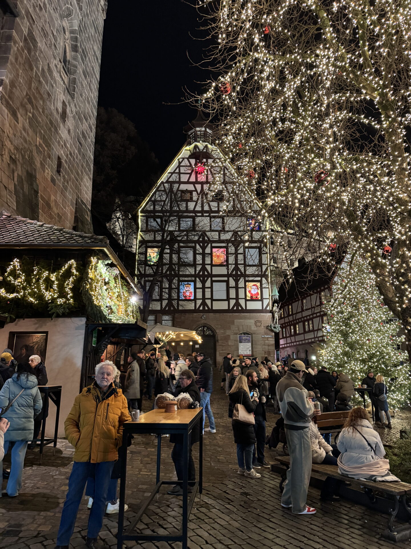 Visitors gather beneath trees covered in thousands of warm white fairy lights beside a traditional half timbered building at night. Colourful illuminated Christmas figures decorate the building windows, whilst a large sparkling Christmas tree stands in the cobbled courtyard.