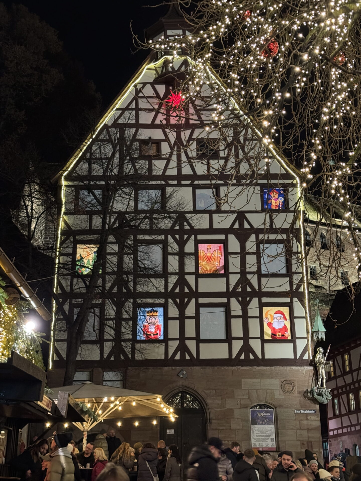A traditional half timbered building illuminated with warm white lights at night, with colourful Christmas characters including Father Christmas displayed in its windows. Visitors gather beneath the building around a warmly lit outdoor drinks stall.