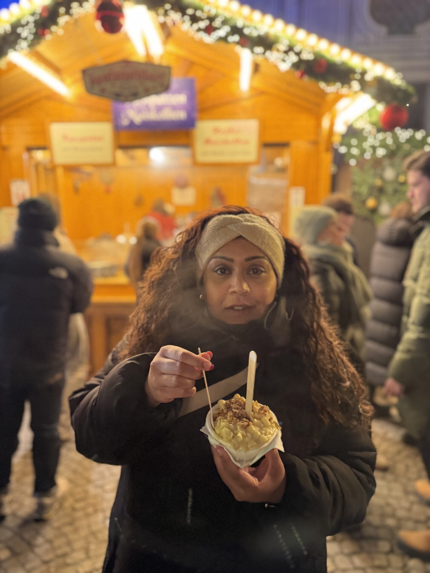 Nishi wearing a cream knitted headband and black winter coat holds a paper tray of creamy Käsespätzle topped with crispy onions. She lifts a wooden fork towards the cheesy German pasta dish while standing in front of a brightly lit Christmas market stall at night.