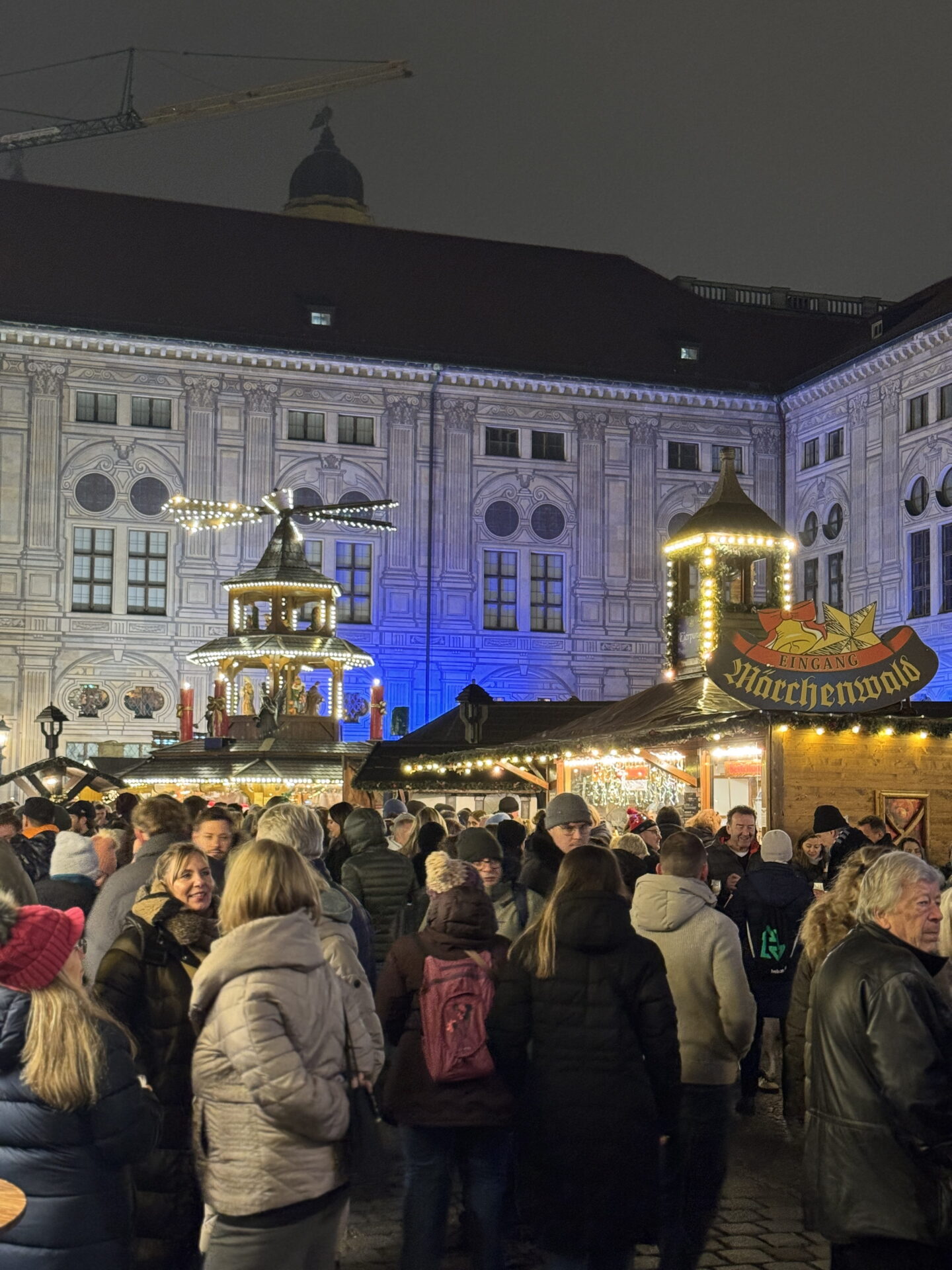 Crowds of visitors in winter coats gather at Munich Medieval Christmas Market at night, surrounded by illuminated wooden stalls and traditional decorative towers. A wooden sign reading "Eingang Märchenwald" hangs above one stall, with a grand historic building glowing blue in the background.