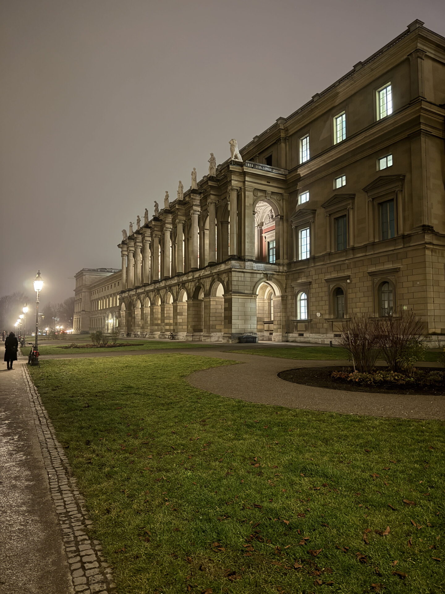 The grand stone façade of Munich Residenz illuminated at night, with rows of arched columns and classical statues lining the upper level. A quiet pathway curves through the lawns beside glowing street lamps beneath a misty evening sky.