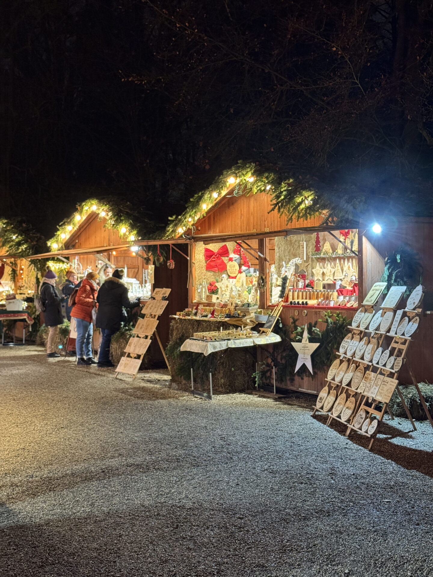 Wooden Christmas market huts decorated with evergreen garlands and warm white fairy lights glow against the dark night sky. Visitors browse handmade Christmas decorations, wooden ornaments and festive gifts displayed outside the stalls.
