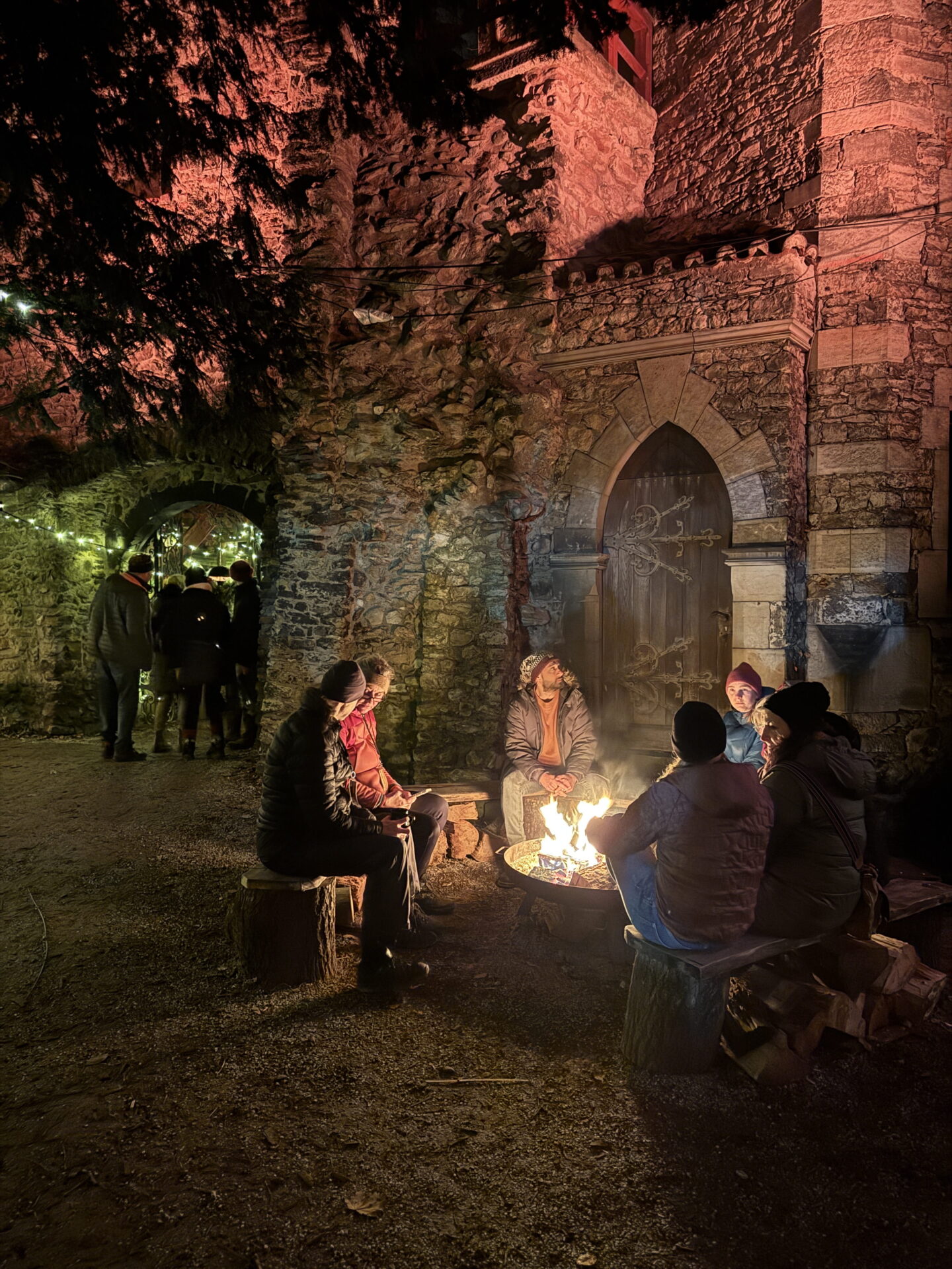 A group of visitors in winter coats and hats sit on rustic wooden benches around a blazing fire bowl at an outdoor Christmas market. The flames illuminate an old stone wall and arched wooden doorway, with more visitors walking through a lit stone passageway.