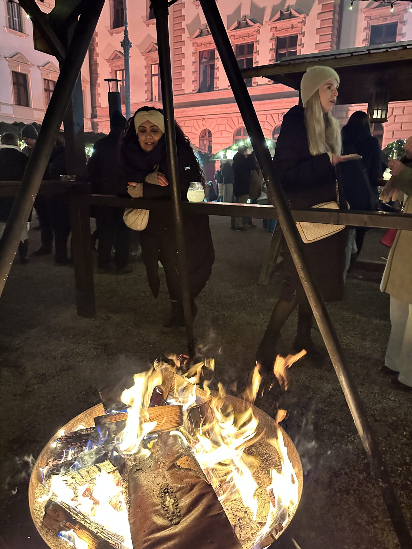 A large wood fire burns beneath a metal shelter at a Christmas market at night. Nishi stands behind a wooden railing, with an ornate historic building illuminated in pink light behind them.