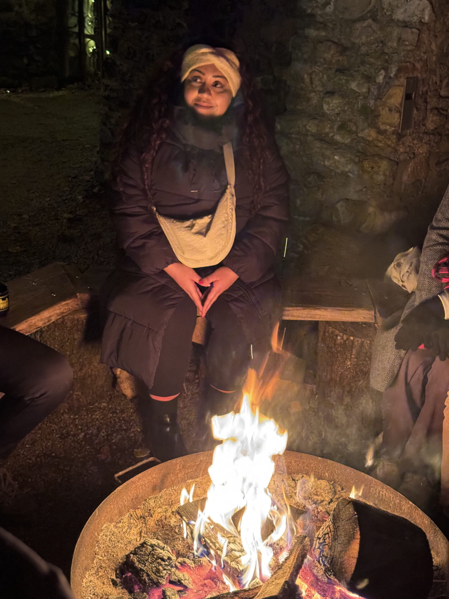 Nishi sits on a wooden bench beside a large fire bowl. Bright flames light her face against the dark stone wall behind her.