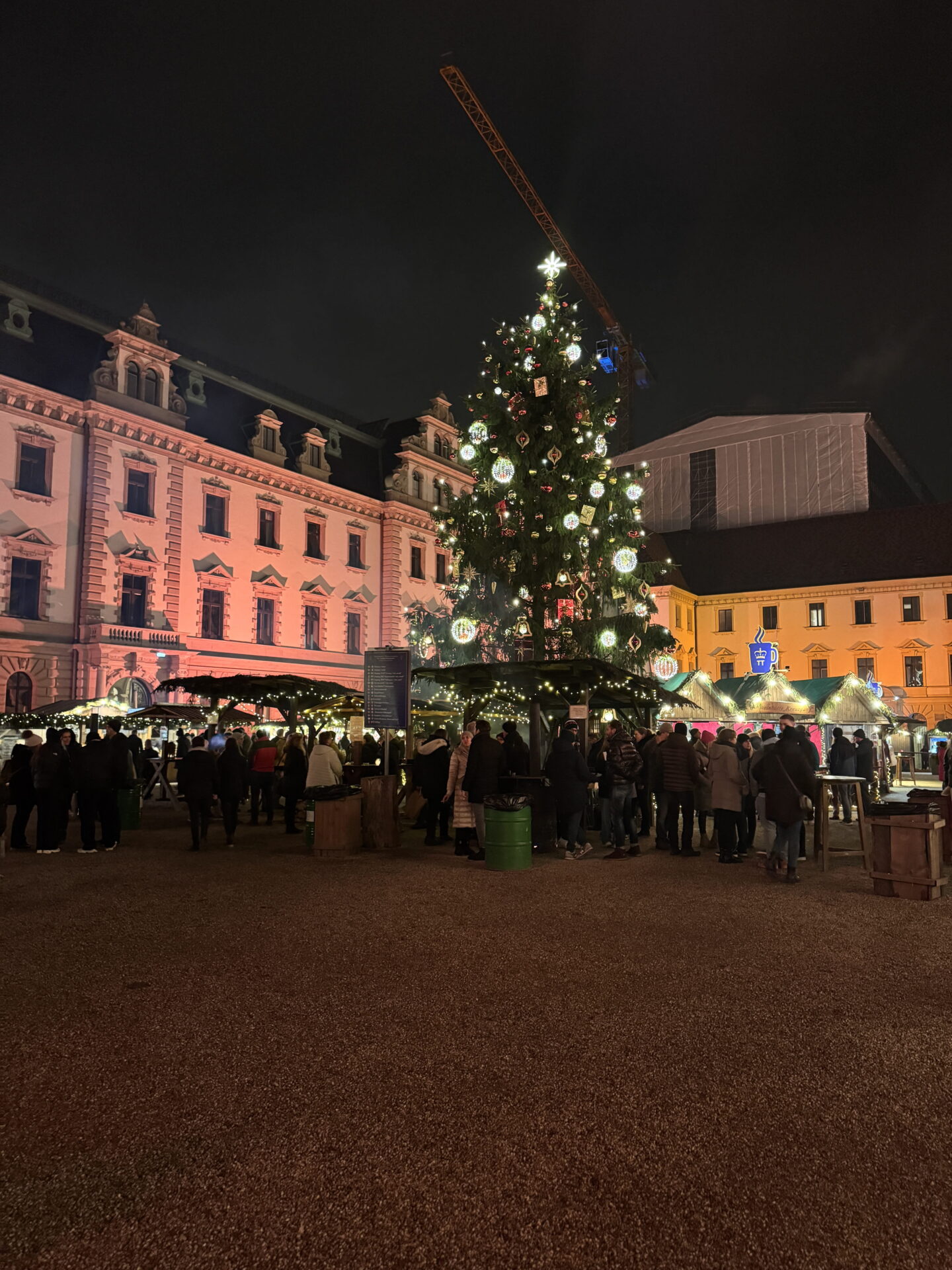 A towering Christmas tree decorated with glowing ornaments and a bright star stands in the centre of a bustling Christmas market at night. Visitors gather beneath the tree and around illuminated wooden stalls, with historic buildings lit in pink and gold.