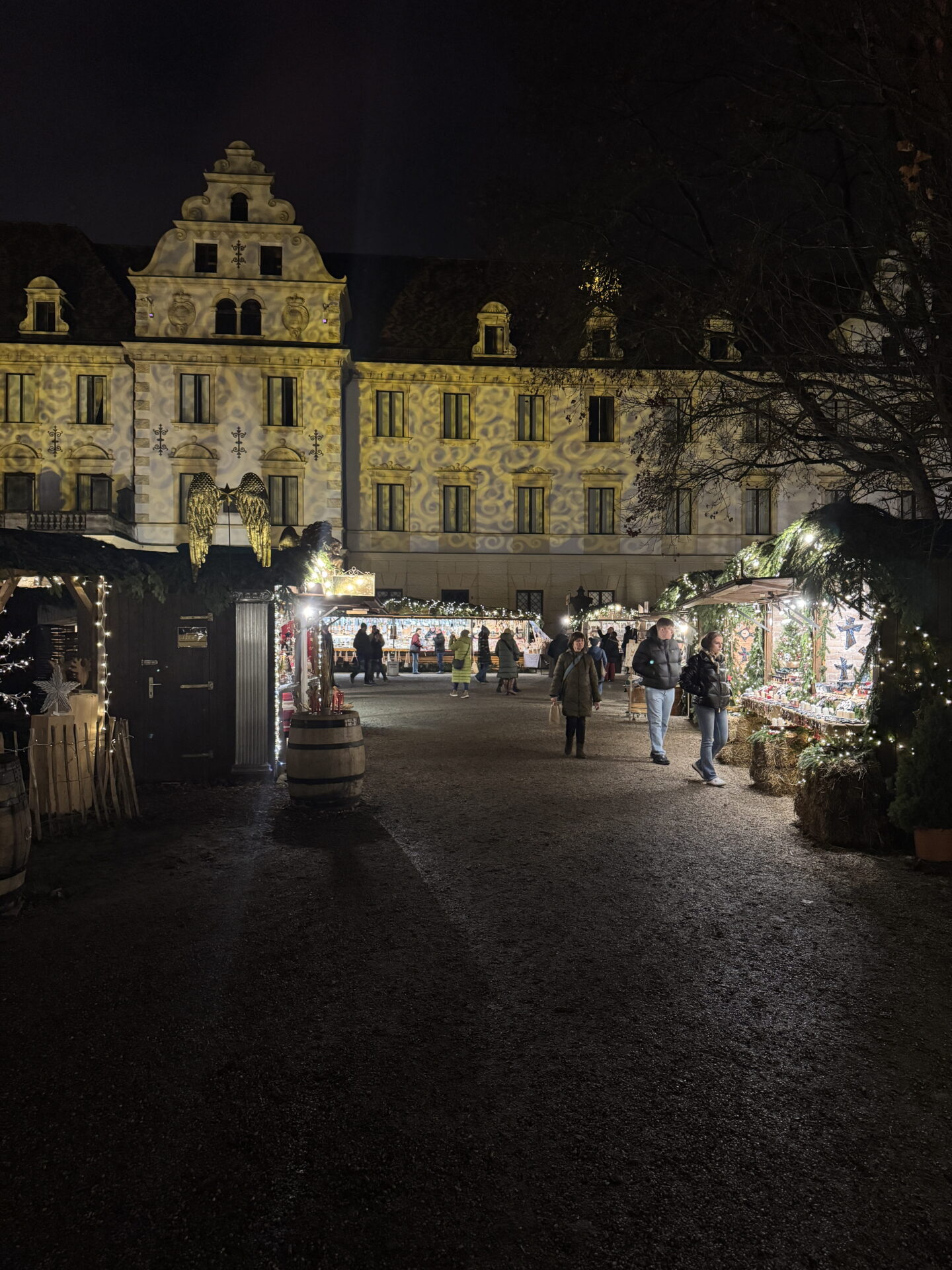 A Christmas market fills a historic courtyard at night, with warmly illuminated wooden stalls displaying festive decorations. Golden patterns are projected across the grand building behind the stalls, while visitors stroll along the gravel pathways.