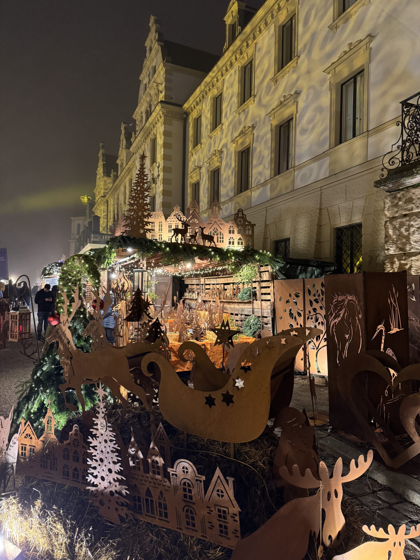 A Christmas market stall displays rustic metal Christmas decorations including reindeer, sleighs, stars, Christmas trees and miniature houses. Warm fairy lights illuminate the handcrafted ornaments against a historic building covered in golden light projections.