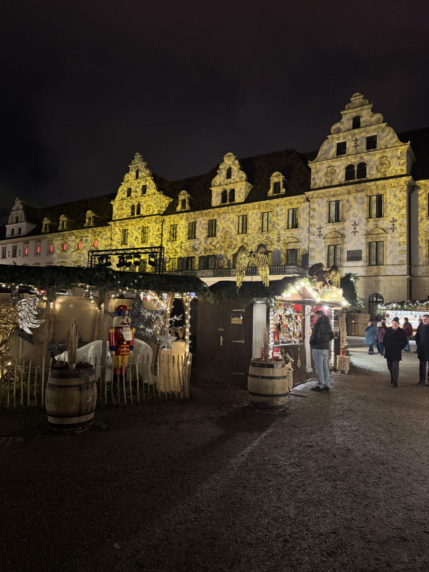 An ornate historic building glows with swirling golden light projections above a Christmas market at night. Wooden stalls decorated with fairy lights, angel ornaments and a colourful nutcracker line the courtyard as visitors browse.