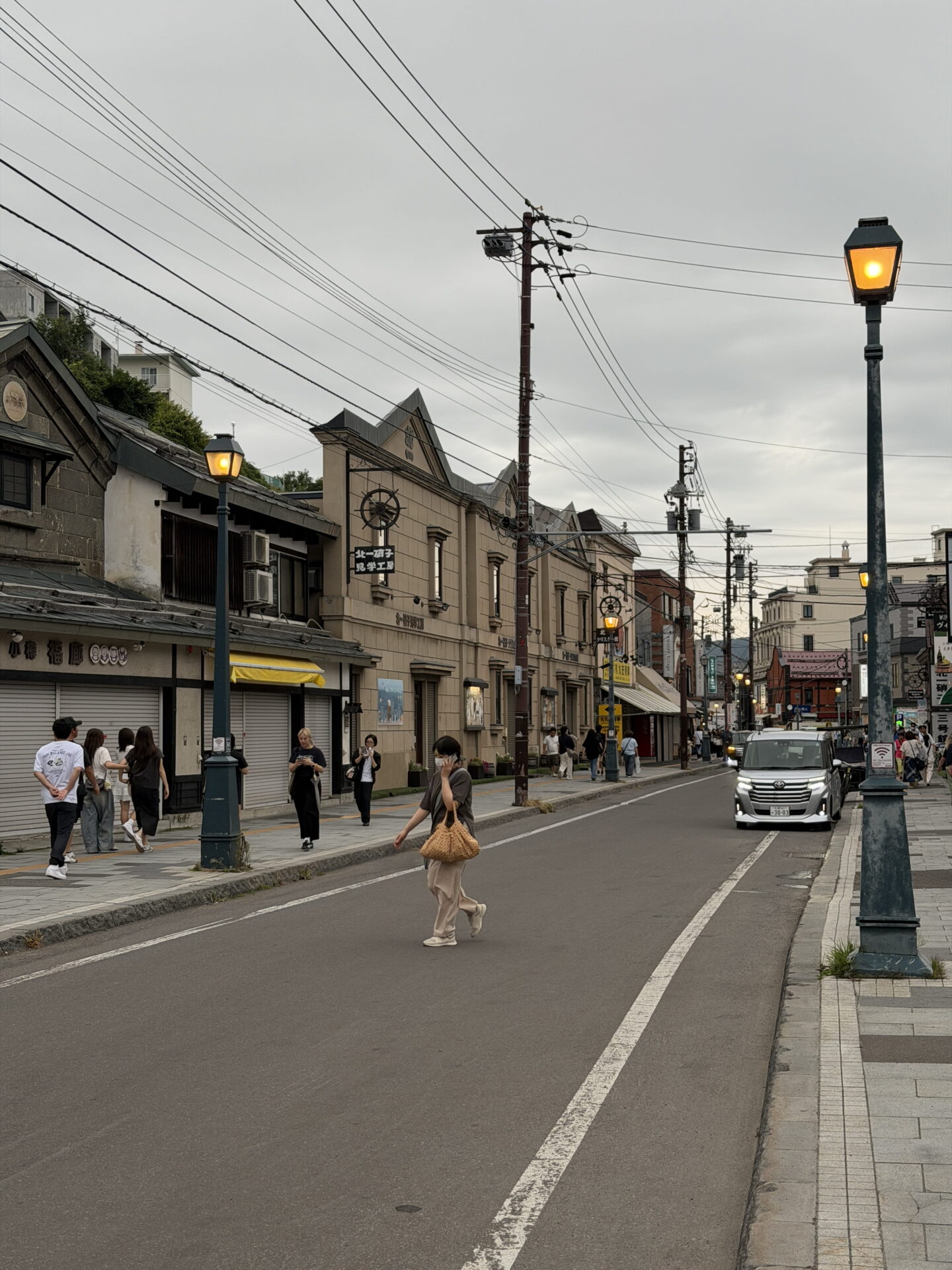 Sakaimachi Street, a historic street in Otaru lined with old stone and brick buildings beneath an overcast evening sky. Traditional style street lamps glow along the pavements while pedestrians walk past shopfronts and a silver vehicle approaches along the road.