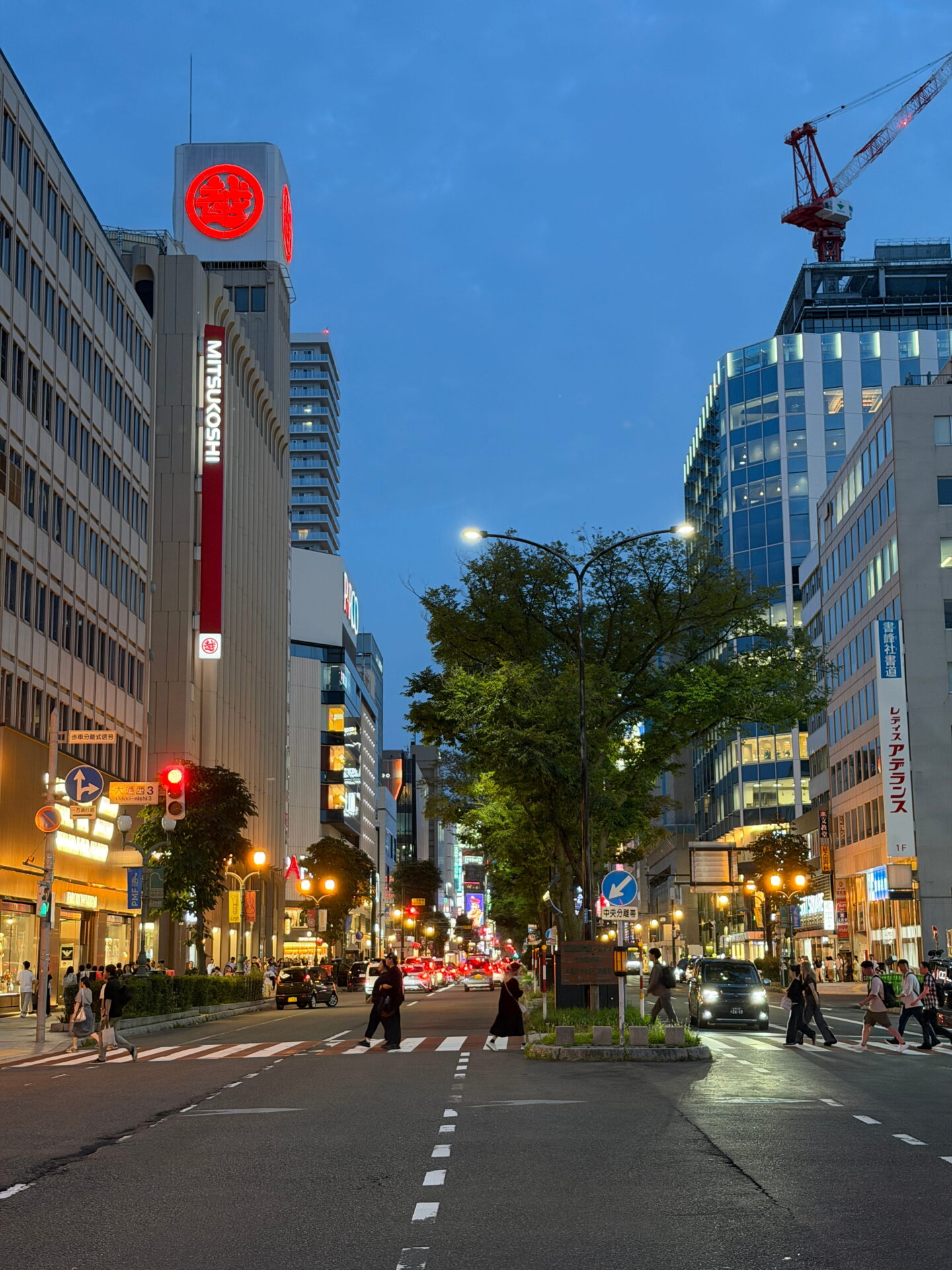 A busy street in central Sapporo at dusk with the illuminated Mitsukoshi department store on the left. Pedestrians cross the road beneath streetlights while cars travel between tall buildings under a deep blue evening sky.