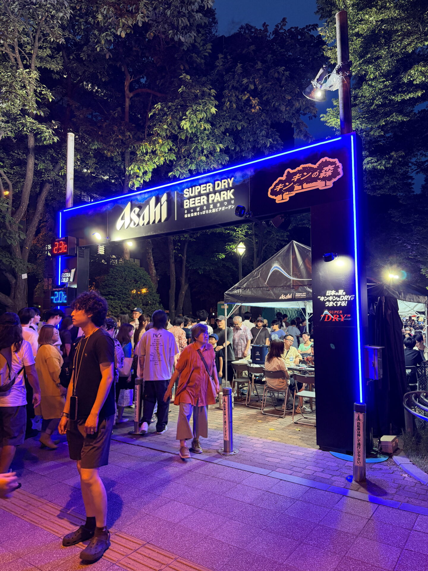 The entrance to the Asahi Super Dry Beer Park in Odori Park in Sapporo at night, outlined in bright blue neon lighting. Crowds gather beneath the illuminated sign while visitors eat and drink at outdoor tables surrounded by trees.