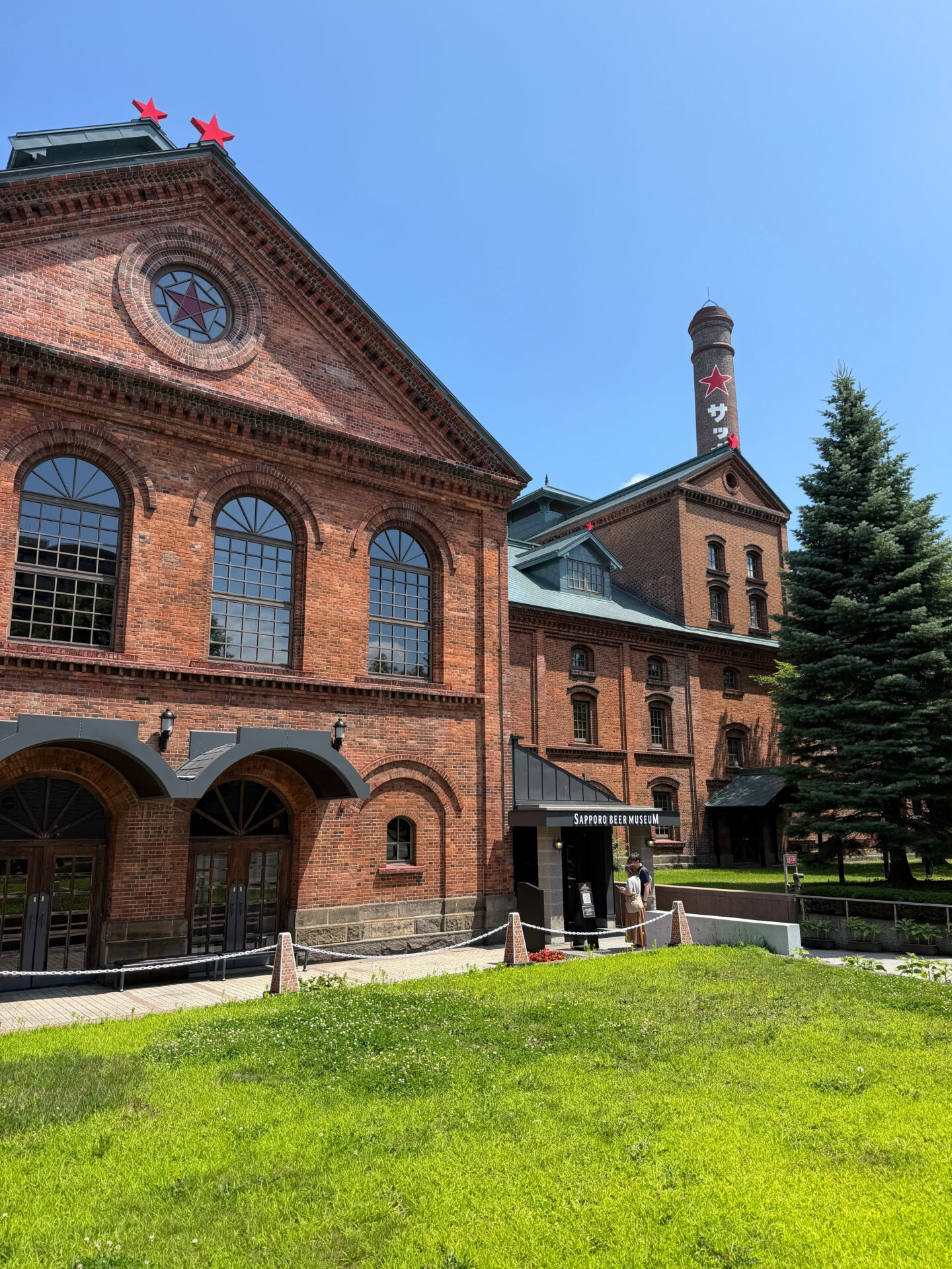 The historic red brick Sapporo Beer Museum on a sunny day, featuring arched windows, green roofs and a tall brick chimney decorated with the Sapporo red star. A small entrance marked Sapporo Beer Museum leads into the former brewery building.