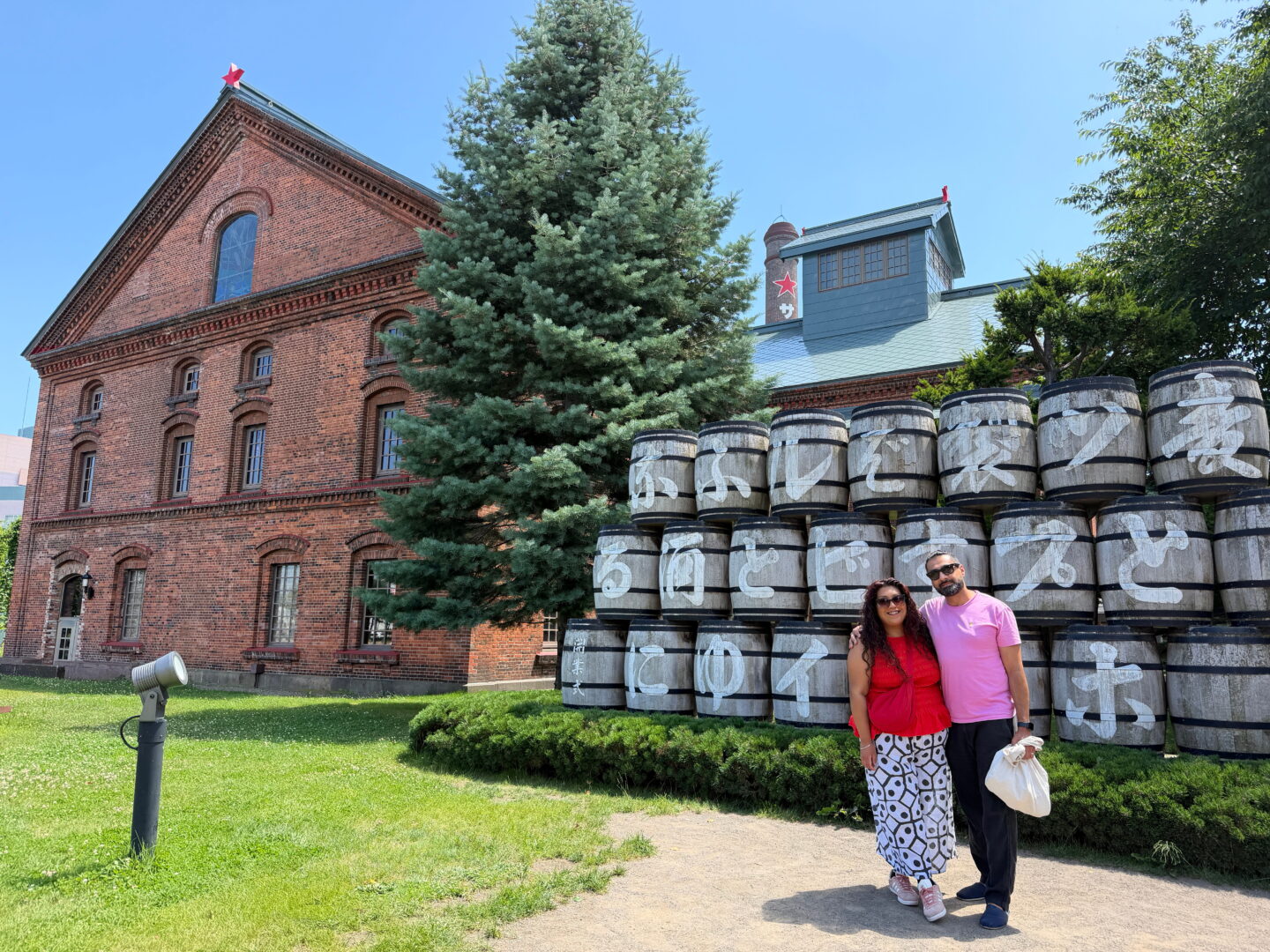 Nishi and her companion pose together in front of a large display of stacked wooden beer barrels at the Sapporo Beer Museum. The couple stand smiling in the sunshine with the historic red brick brewery buildings and a tall evergreen tree behind them.