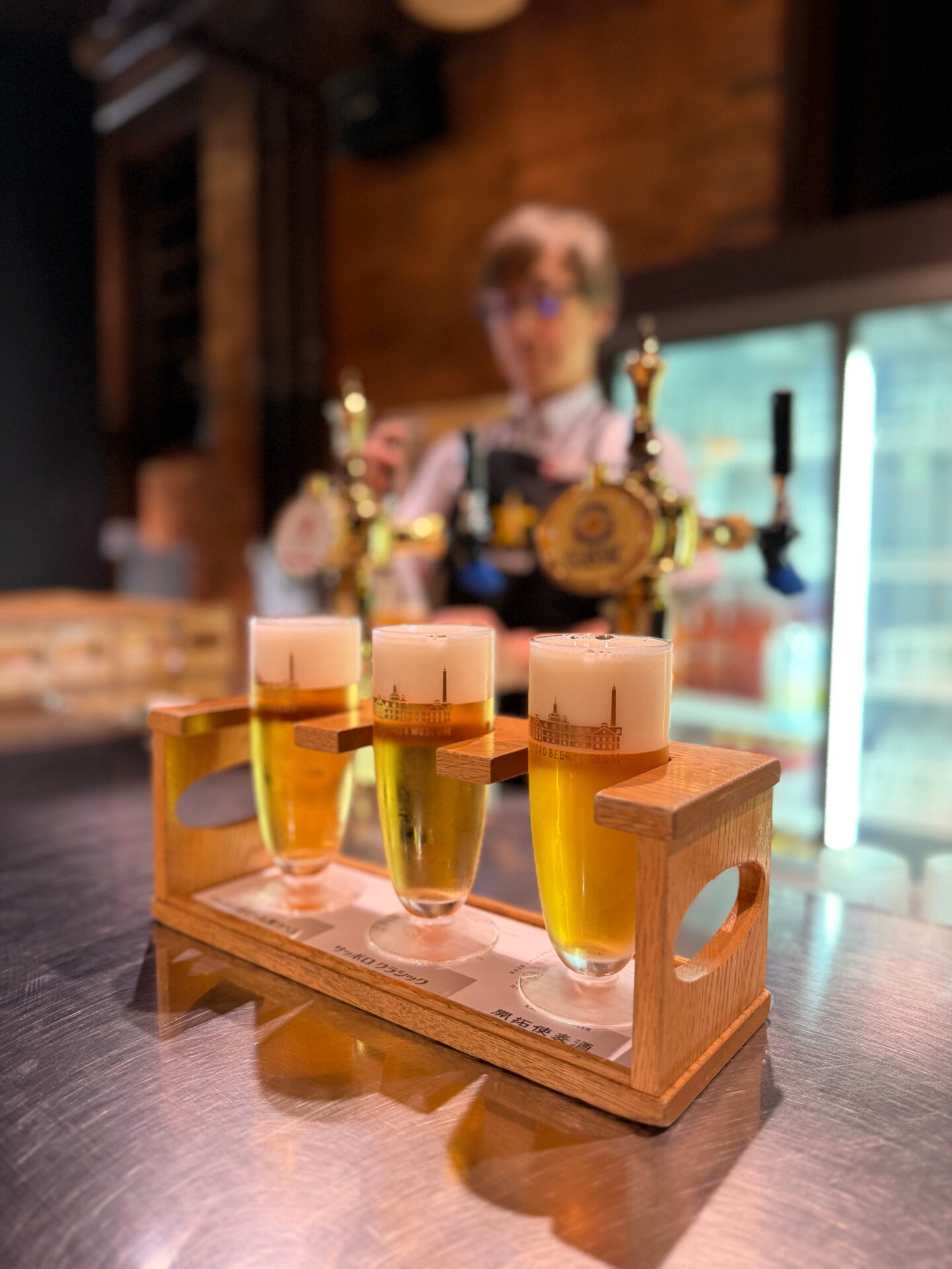 Three small glasses of golden Sapporo beer topped with thick white foam are arranged in a wooden tasting flight at the Sapporo Beer Museum. Brass beer taps and a member of staff are softly blurred in the background.