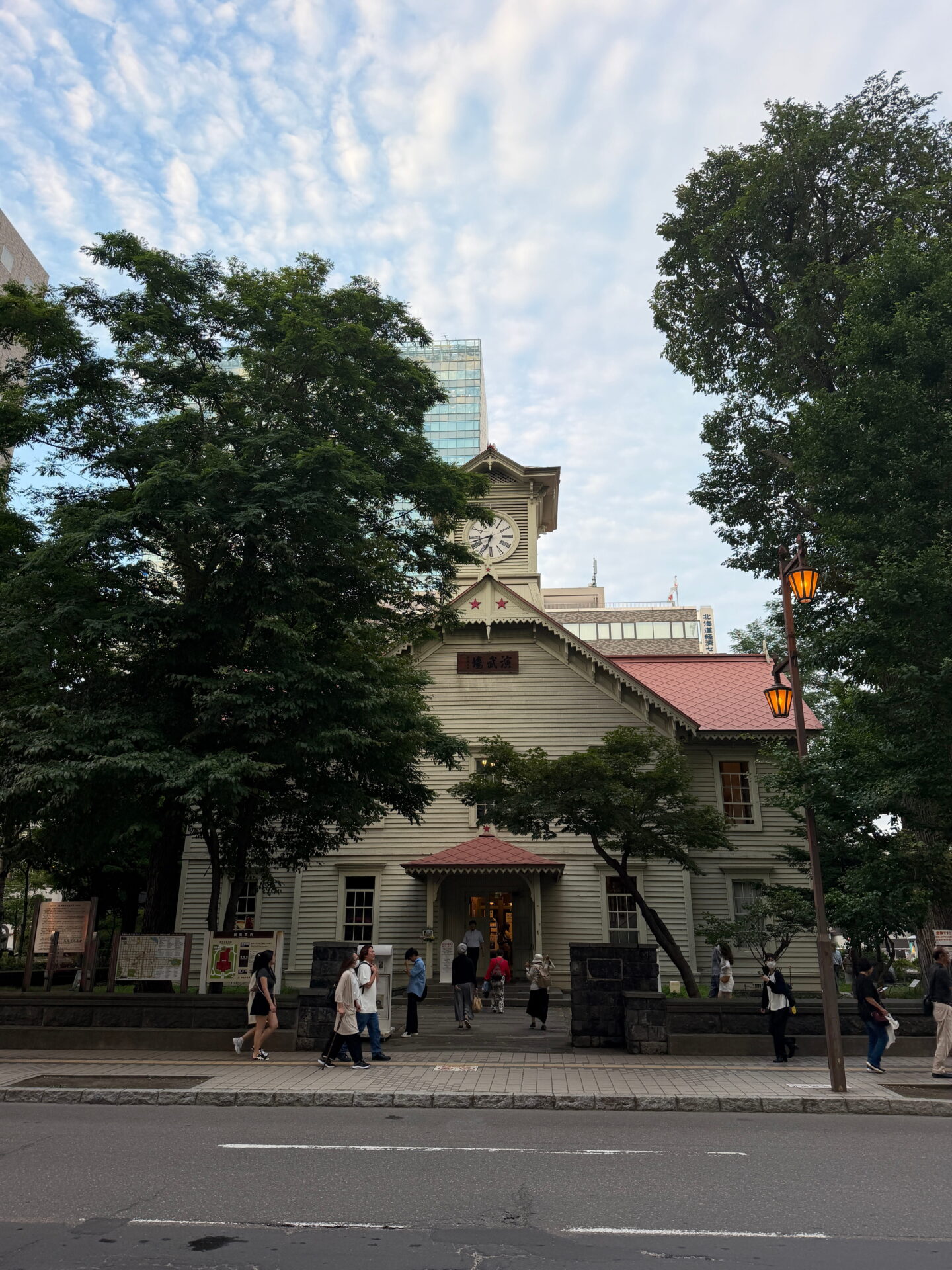 The historic Sapporo Clock Tower in central Sapporo, a pale wooden building with a red roof, triangular gables and a white clock face. Visitors walk past the entrance while large leafy trees frame the building against a cloudy evening sky.