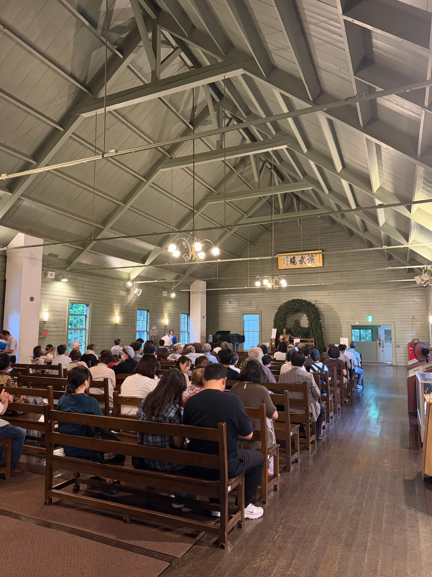 Visitors sit on rows of wooden benches inside the historic Sapporo Clock Tower, beneath a high ceiling of exposed white painted timber beams. Chandeliers hang overhead while people face a small presentation area at the front of the hall.