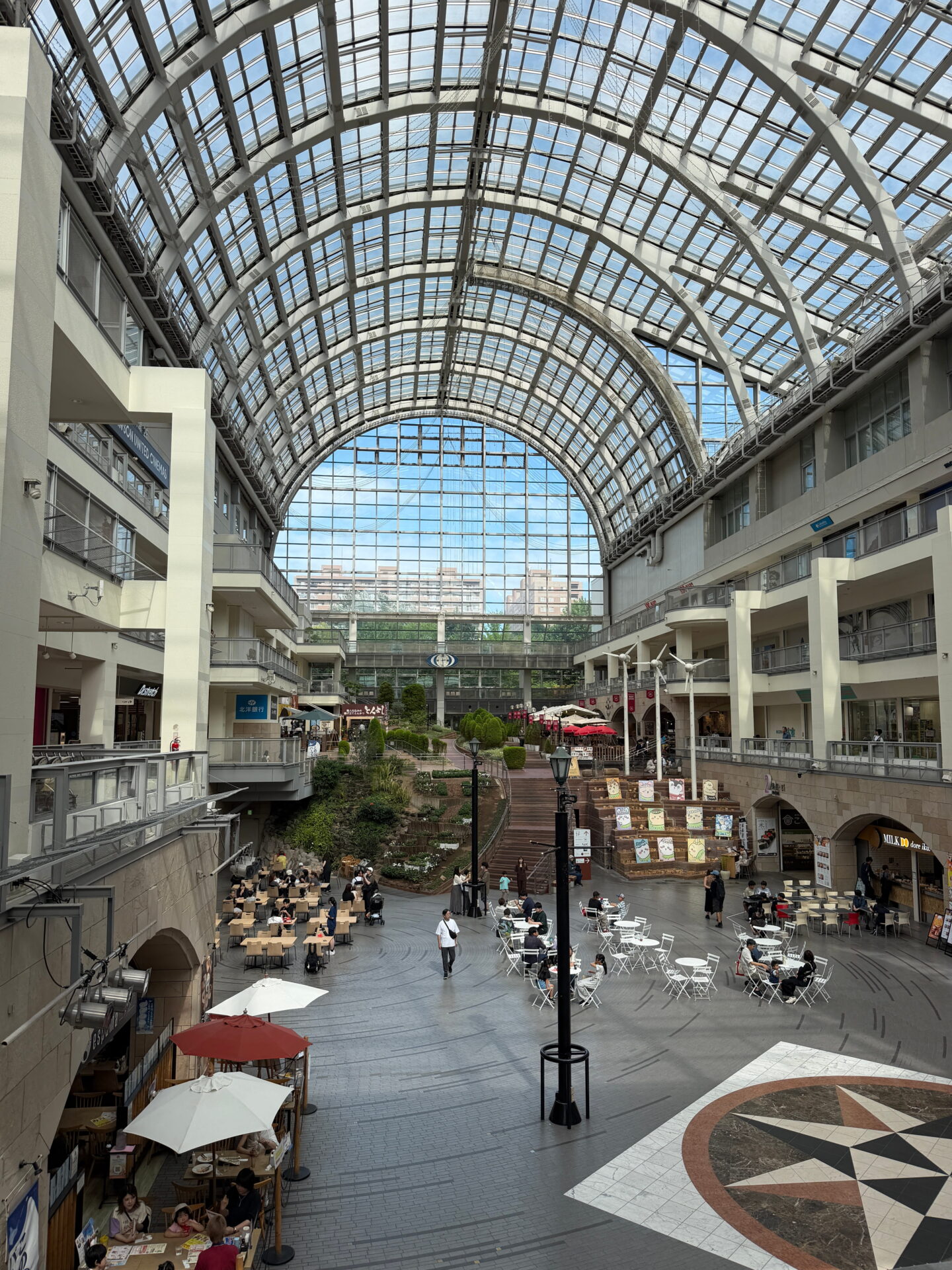 The spacious indoor atrium of Sapporo Factory shopping centre, featuring a huge arched glass roof, restaurants, shops and outdoor style café seating. A landscaped garden and wide staircase occupy the centre of the bright multi level shopping complex.