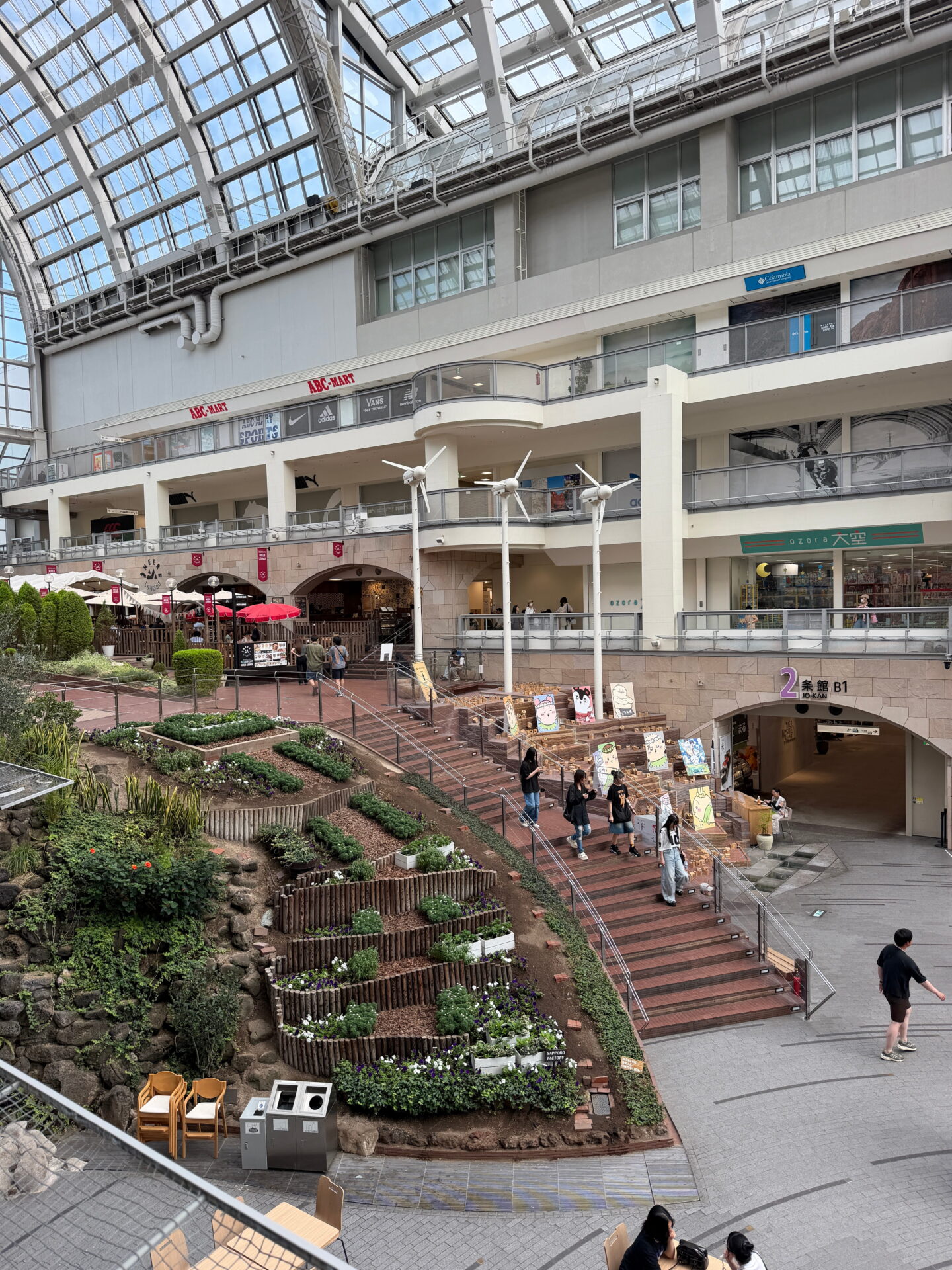 A closer view of the Sapporo Factory shopping centre atrium with terraced flowerbeds, wide brick steps and small decorative wind turbines. Shoppers walk between restaurants and shops across several levels beneath the enormous glass roof.