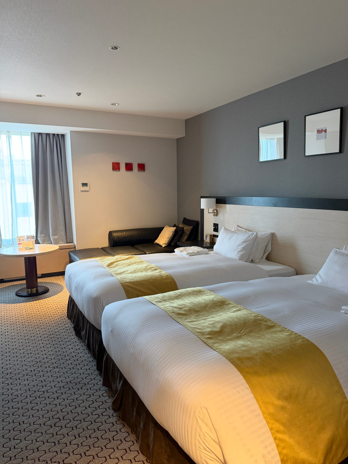 A spacious hotel room in Sapporo with two double beds dressed in white linen and gold runners. A black leather sofa, small round table and large curtained window complete the room.