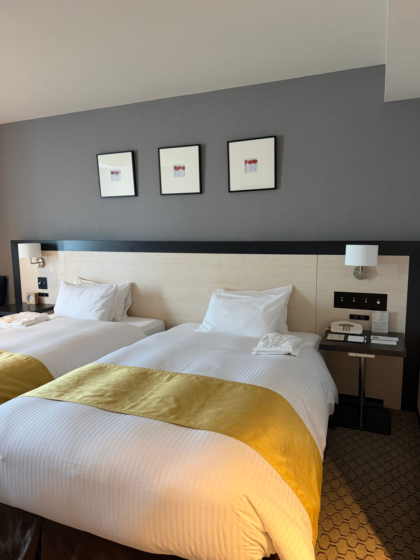 A bright twin bedroom in a Sapporo hotel with two neatly made single beds dressed in white striped bedding and mustard yellow runners. Matching bedside lamps, folded towels and three framed floral prints complete the modern room.