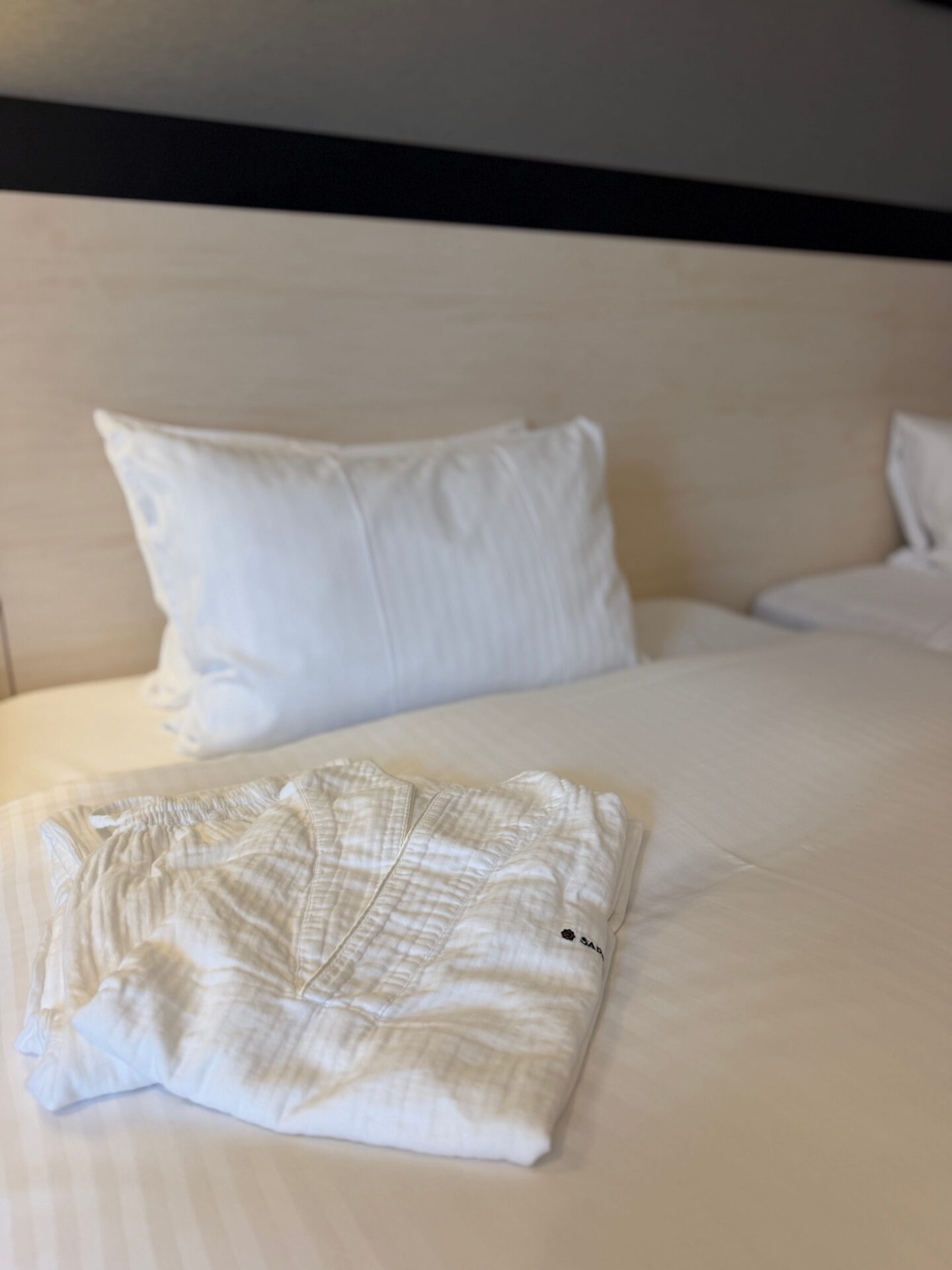 A neatly folded pair of white textured hotel pyjamas rests on a bed with crisp white bedding and a large pillow. A light wooden headboard completes the simple hotel room setting in Sapporo.