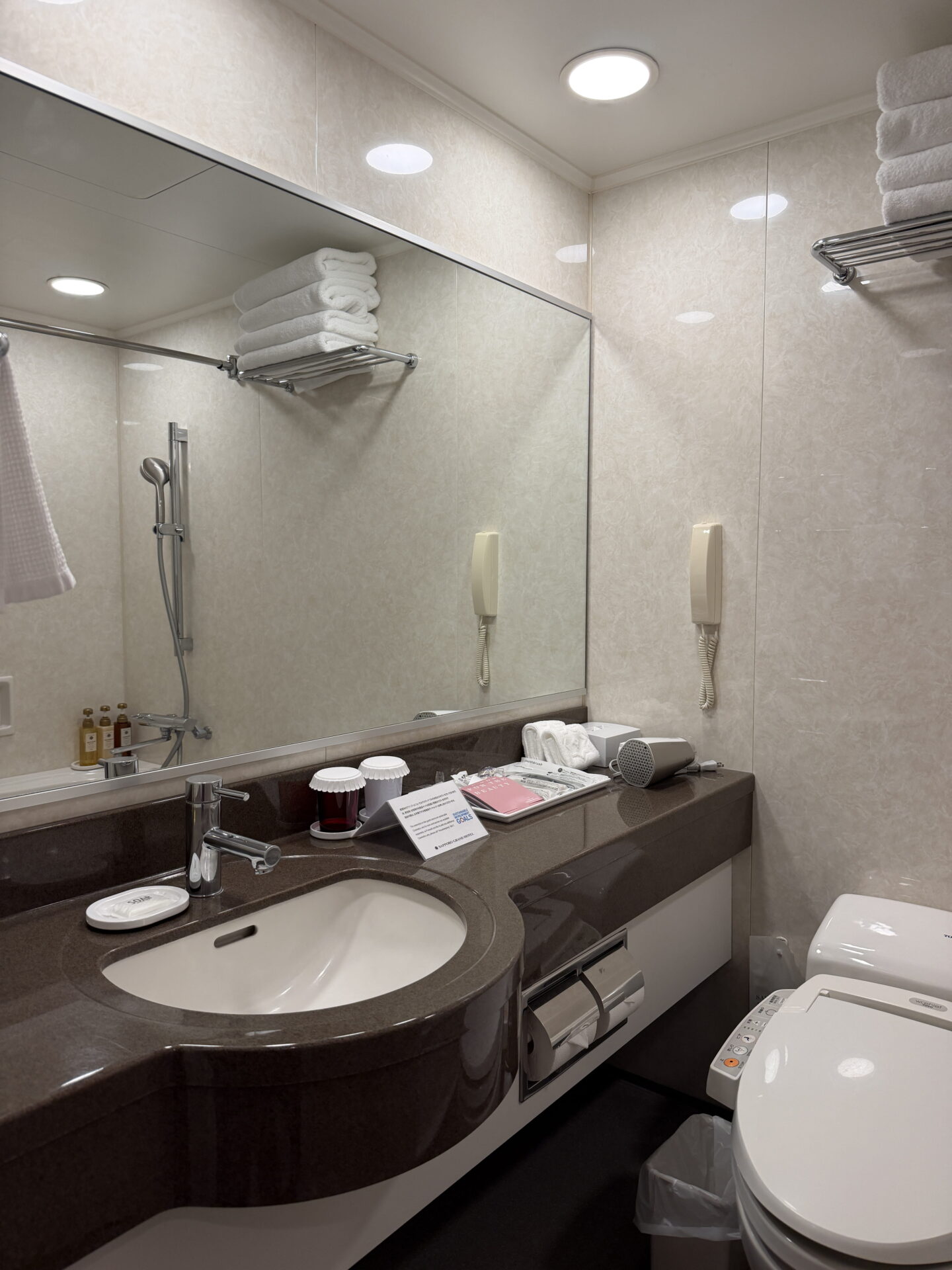 A bathroom at the Sapporo Grand Hotel featuring a dark stone countertop with an inset white sink and a large wall mirror. A Japanese bidet toilet sits beside the vanity with a hairdryer and toiletries provided and neatly folded white towels above the shower.