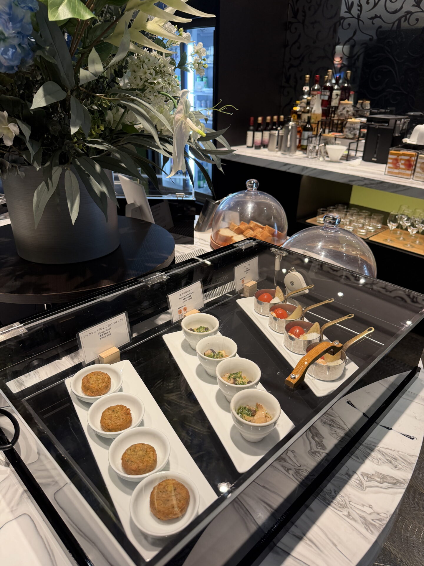 A selection of canapés and small appetisers displayed in white bowls and miniature pans beneath a glass cover at the Sapporo Grand Hotel Club Lounge. A large arrangement of white flowers sits beside the buffet with drinks and glassware visible behind it.