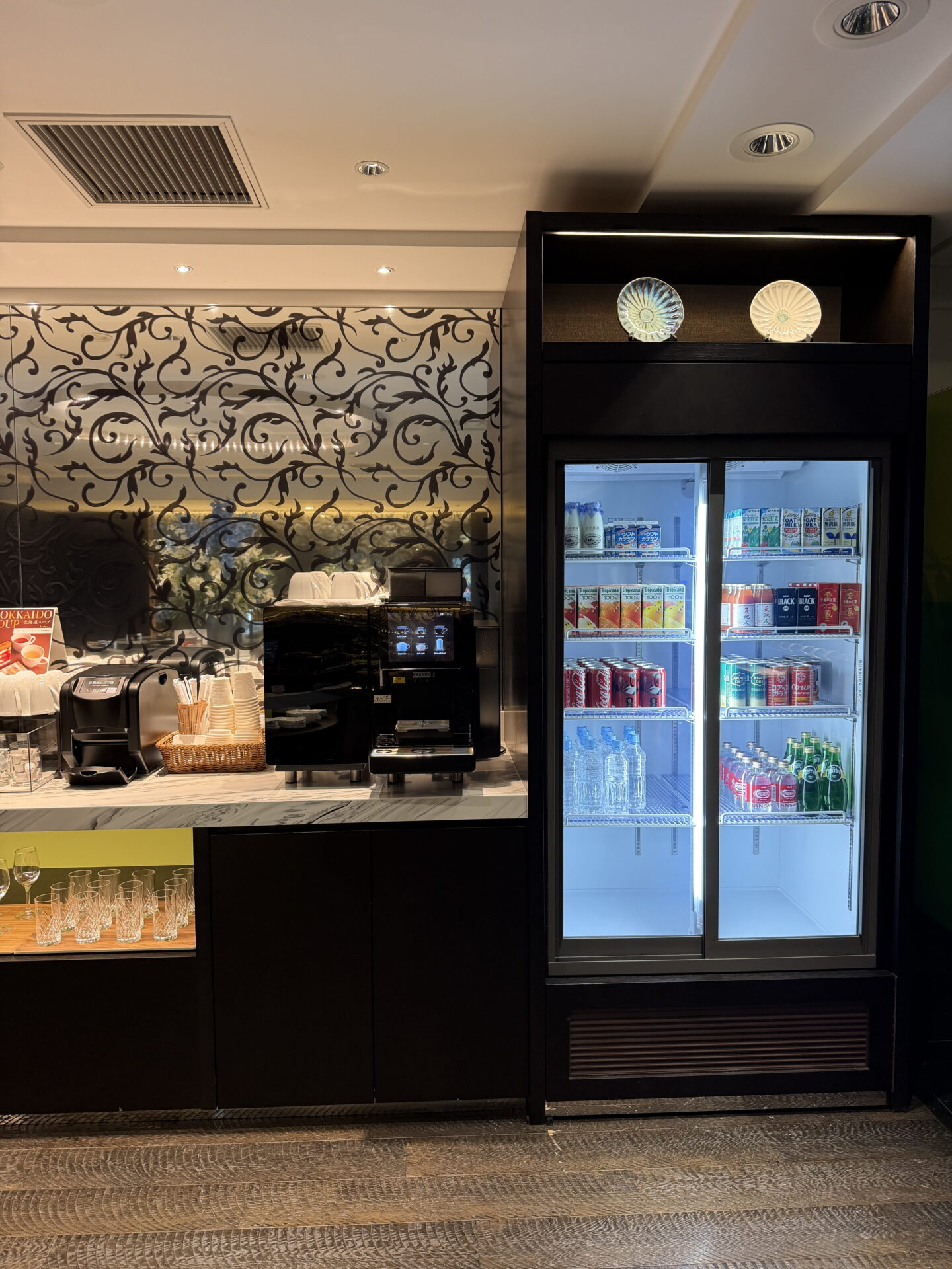 A self service drinks station at the Sapporo Grand Hotel Club Lounge with coffee machines and a glass fronted fridge stocked with bottled water, fruit juices, soft drinks and milk. Cups and glasses are neatly arranged beside a decorative black floral patterned wall.