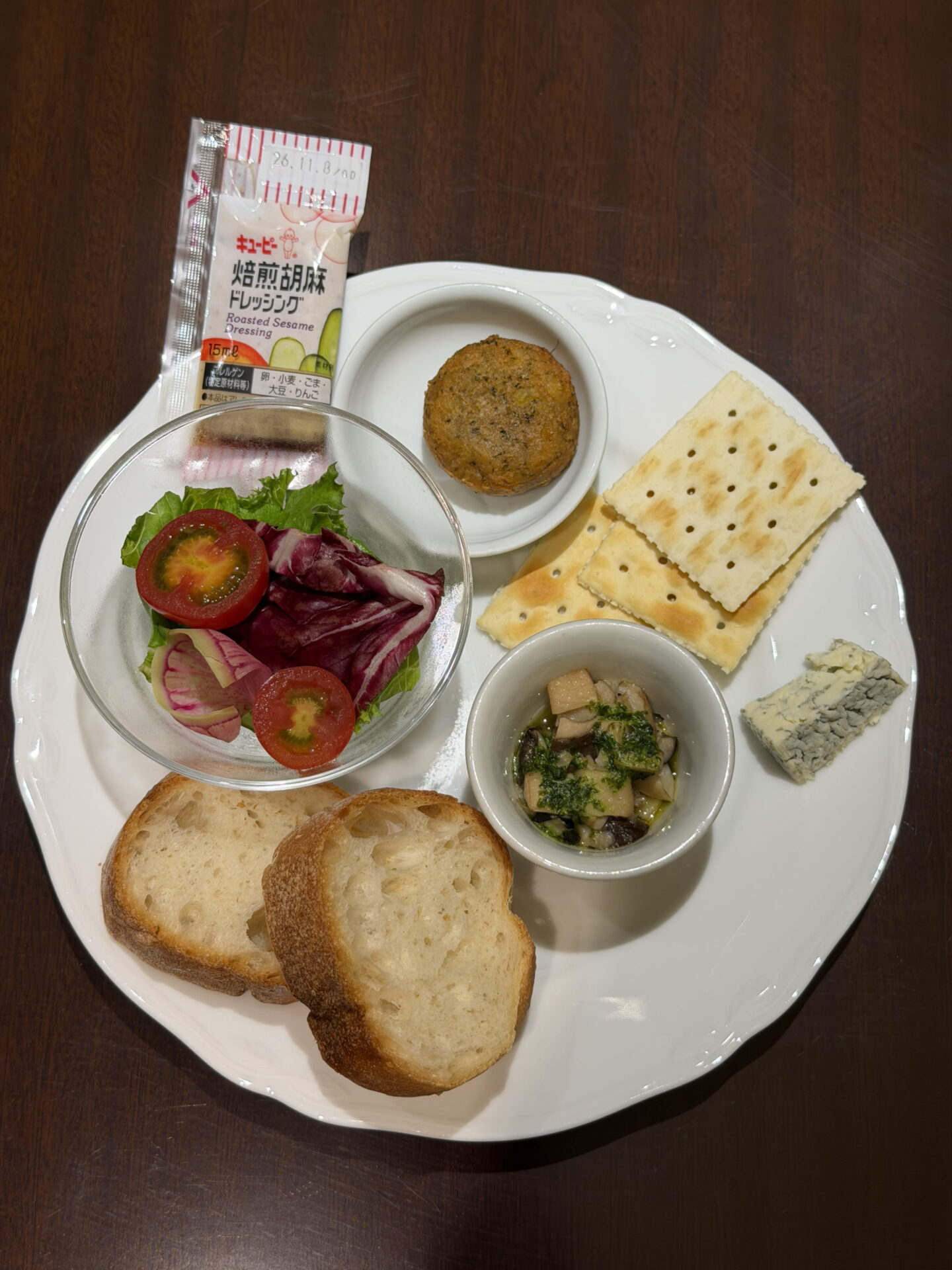 A white plate of appetisers from the Sapporo Grand Hotel Club Lounge featuring slices of crusty bread, crackers, blue cheese, a small savoury croquette and mushrooms in herb dressing. A glass bowl contains mixed salad leaves and cherry tomatoes with a sachet labelled "Roasted Sesame Dressing" alongside.