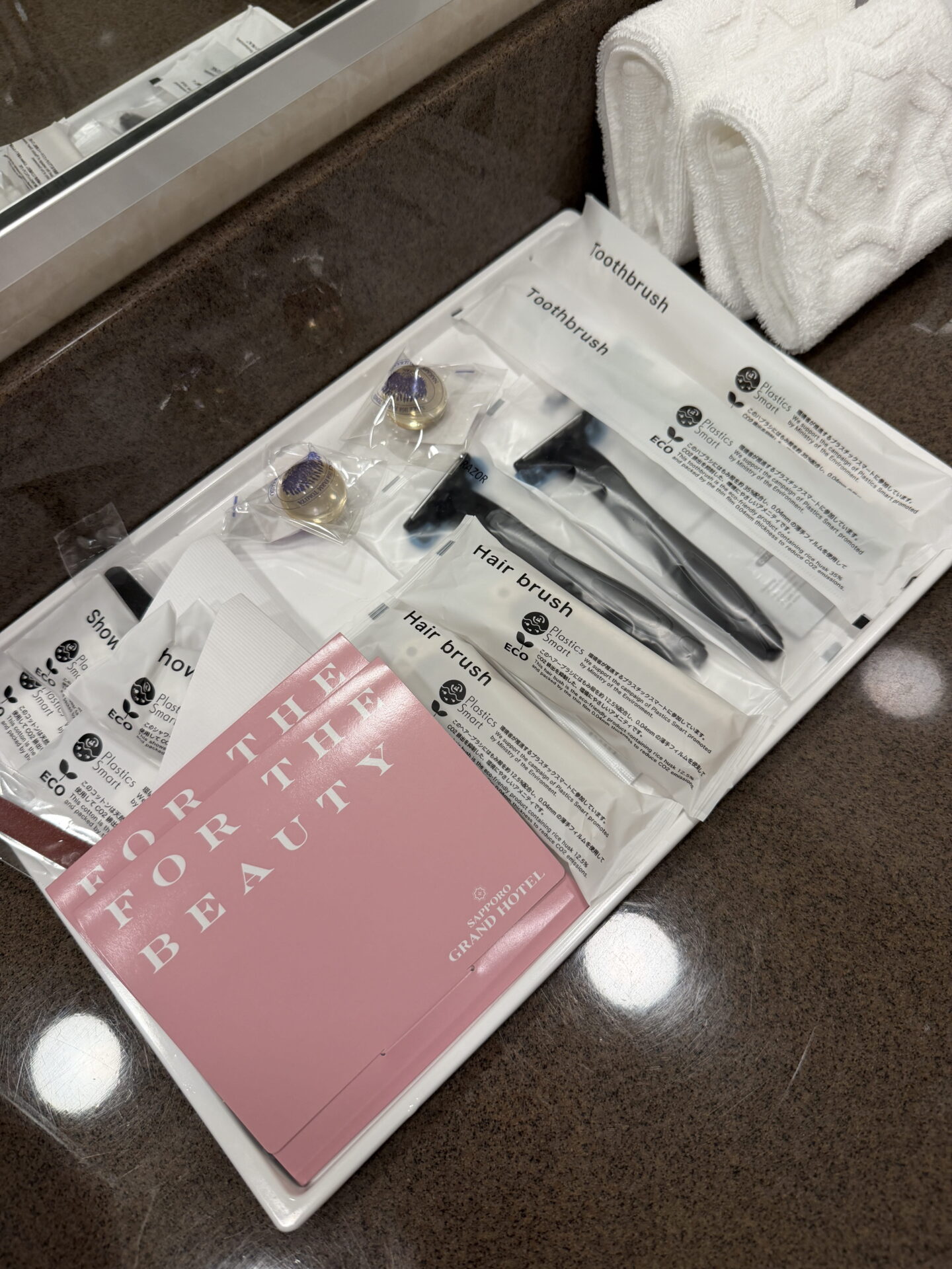 Complimentary bathroom amenities at the Sapporo Grand Hotel arranged on a white tray beside folded towels. Individually wrapped toothbrushes, hairbrushes, shower caps and other toiletries sit alongside pink packets labelled "FOR THE BEAUTY" and "SAPPORO GRAND HOTEL".