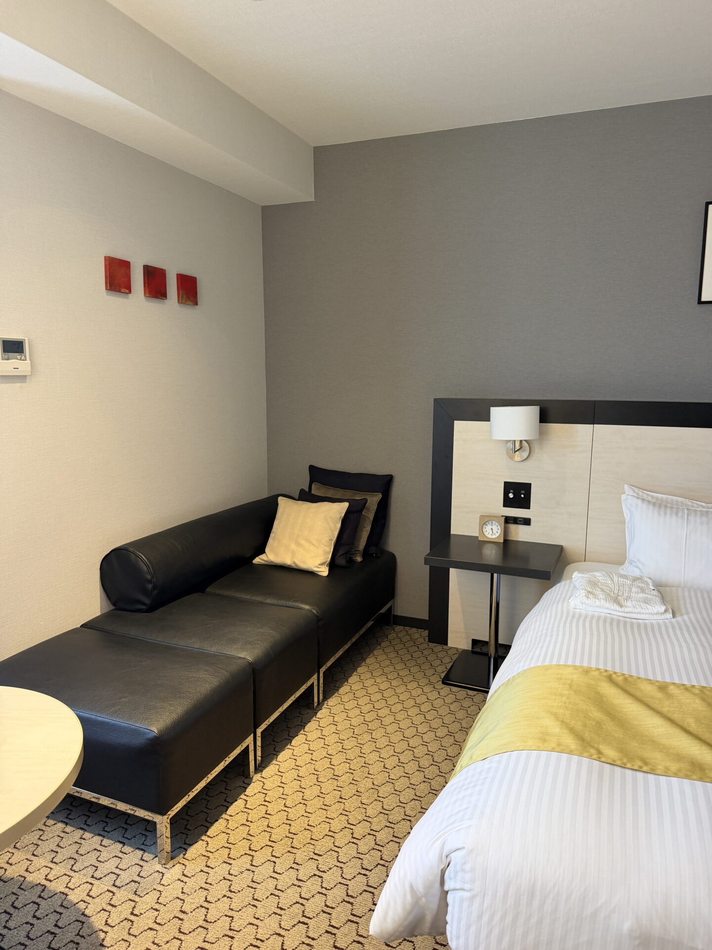 A guest room at the Sapporo Grand Hotel featuring a black leather chaise longue with cream and dark cushions beside a bed dressed in white striped linen and a gold runner. Grey walls, a patterned carpet, a bedside clock and a wall mounted reading light complete the room.