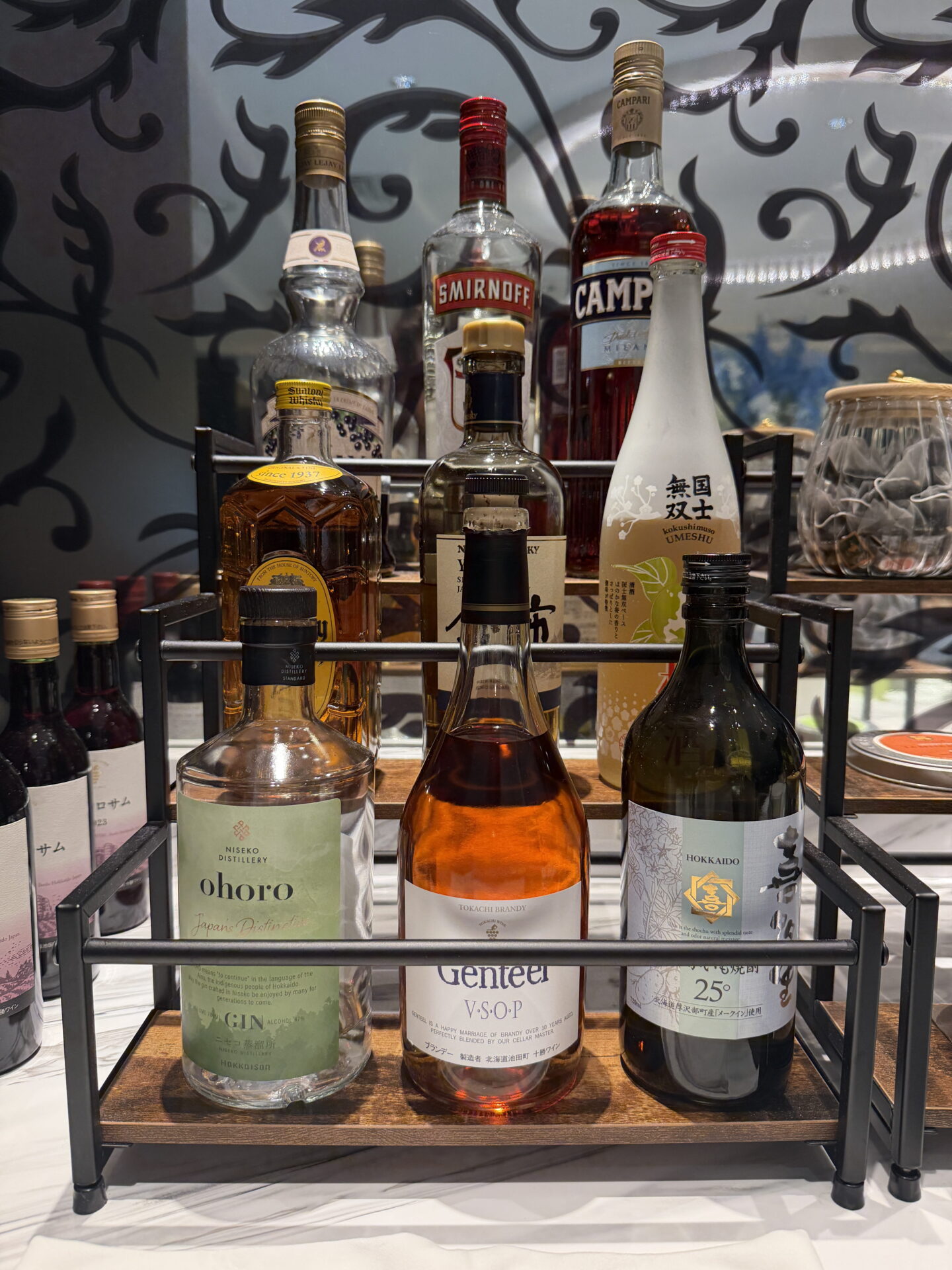 A selection of Japanese and international spirits displayed on a tiered wooden and metal stand at the Sapporo Grand Hotel Club Lounge. Bottles include "ohoro" gin from Niseko Distillery, "Gentéer VSOP" Tokachi brandy, Hokkaido shochu, Smirnoff vodka and Campari.