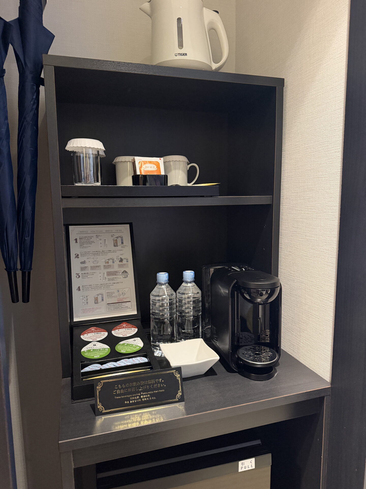 In room tea and coffee facilities at the Sapporo Grand Hotel arranged on a dark wooden shelving unit. A capsule coffee machine, coffee pods, bottled water, mugs, glasses and a white Tiger electric kettle are provided alongside two folded umbrellas and a small fridge below.