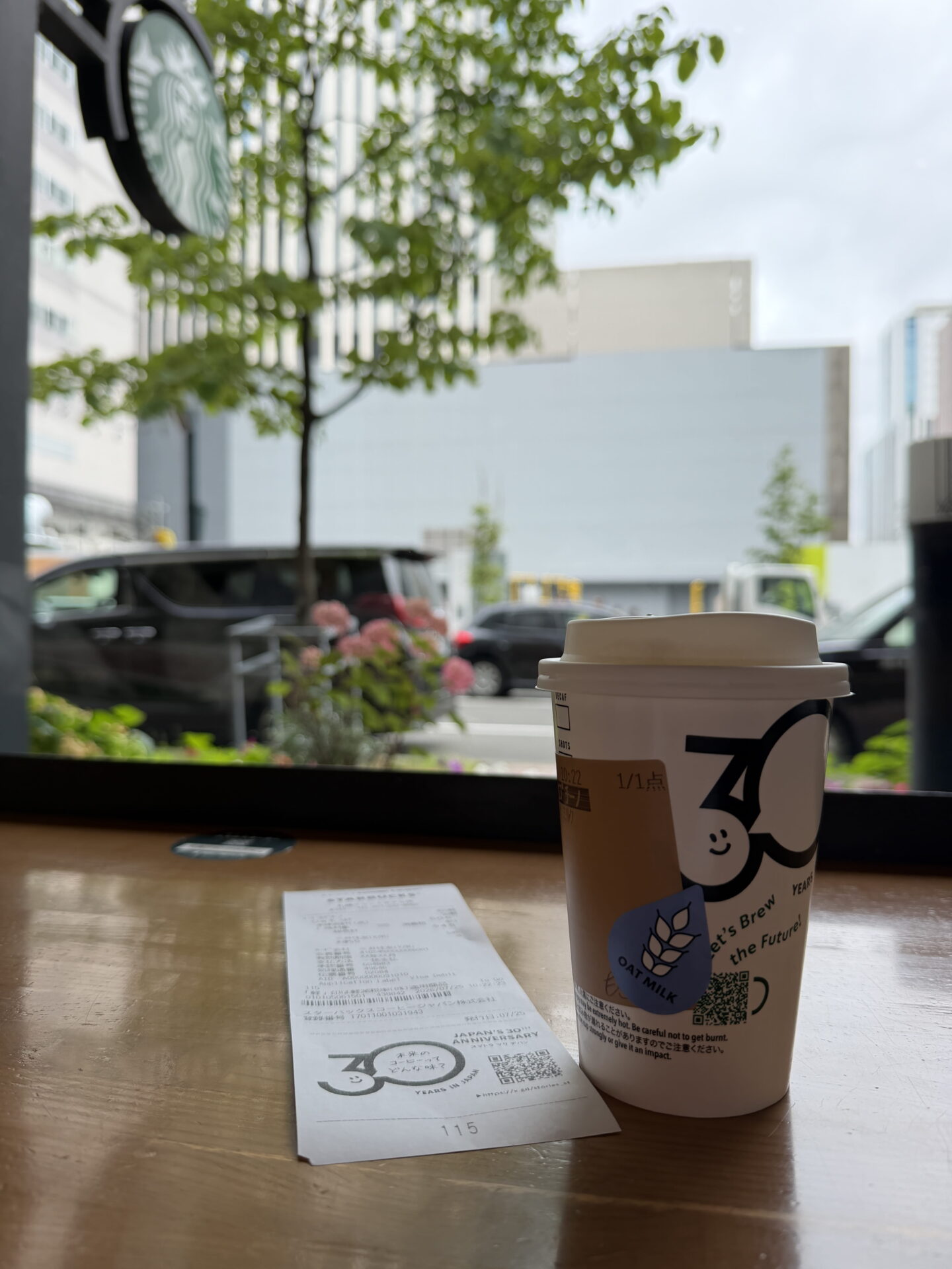 A takeaway Starbucks oat milk coffee in a white anniversary cup featuring the number "30" and the words "Let's Brew the Future" sits beside a receipt on a wooden table. Through the café window in Sapporo are leafy trees, pink flowers, parked cars and city buildings.