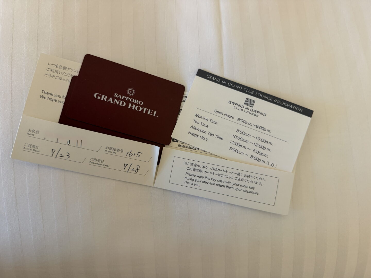 A burgundy room key holder labelled "SAPPORO GRAND HOTEL" sits on white hotel bedding beside an information card for the "GRAND IN GRAND CLUB LOUNGE". The card lists opening hours from 8.00am to 9.00pm with morning time, tea time, afternoon tea time and happy hour sessions.