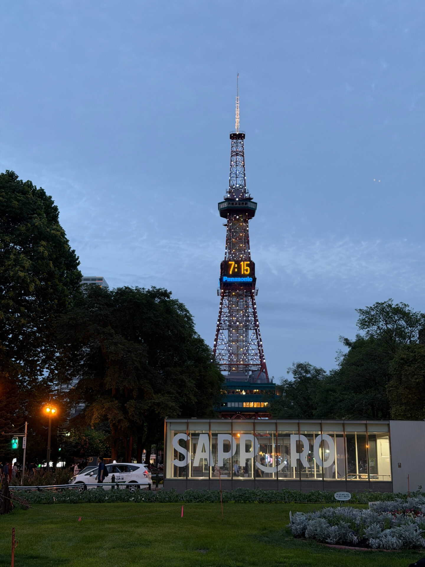 Sapporo TV Tower illuminated against a pale blue evening sky with its digital clock displaying 7.15 and the Panasonic logo beneath it. The glass Sapporo sign stands in the foreground surrounded by trees and greenery.