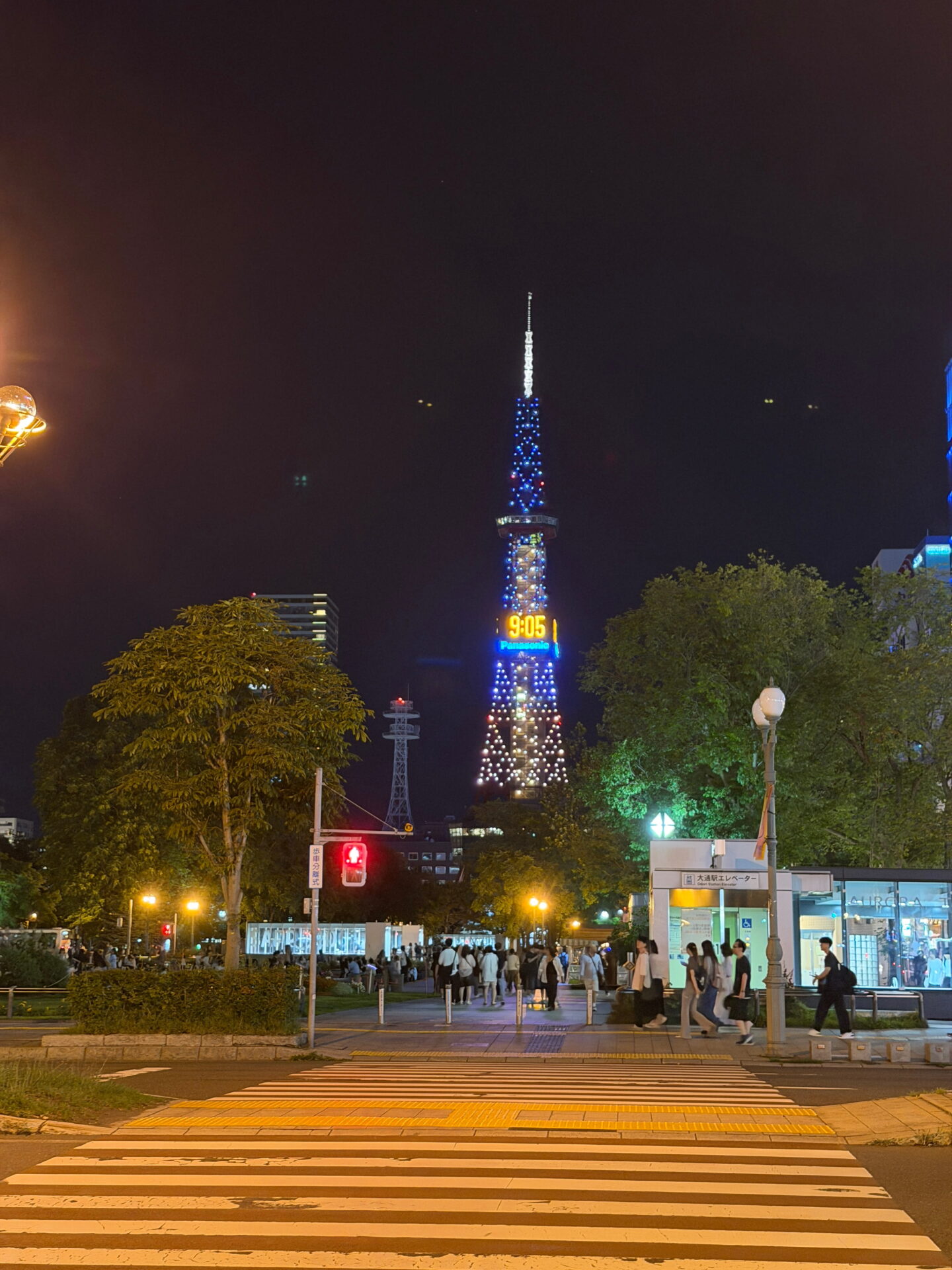 Sapporo TV Tower glowing with blue and white lights against the dark night sky with its digital clock displaying 9.05. A pedestrian crossing leads towards the tower with people walking through the streets near Odori Park.