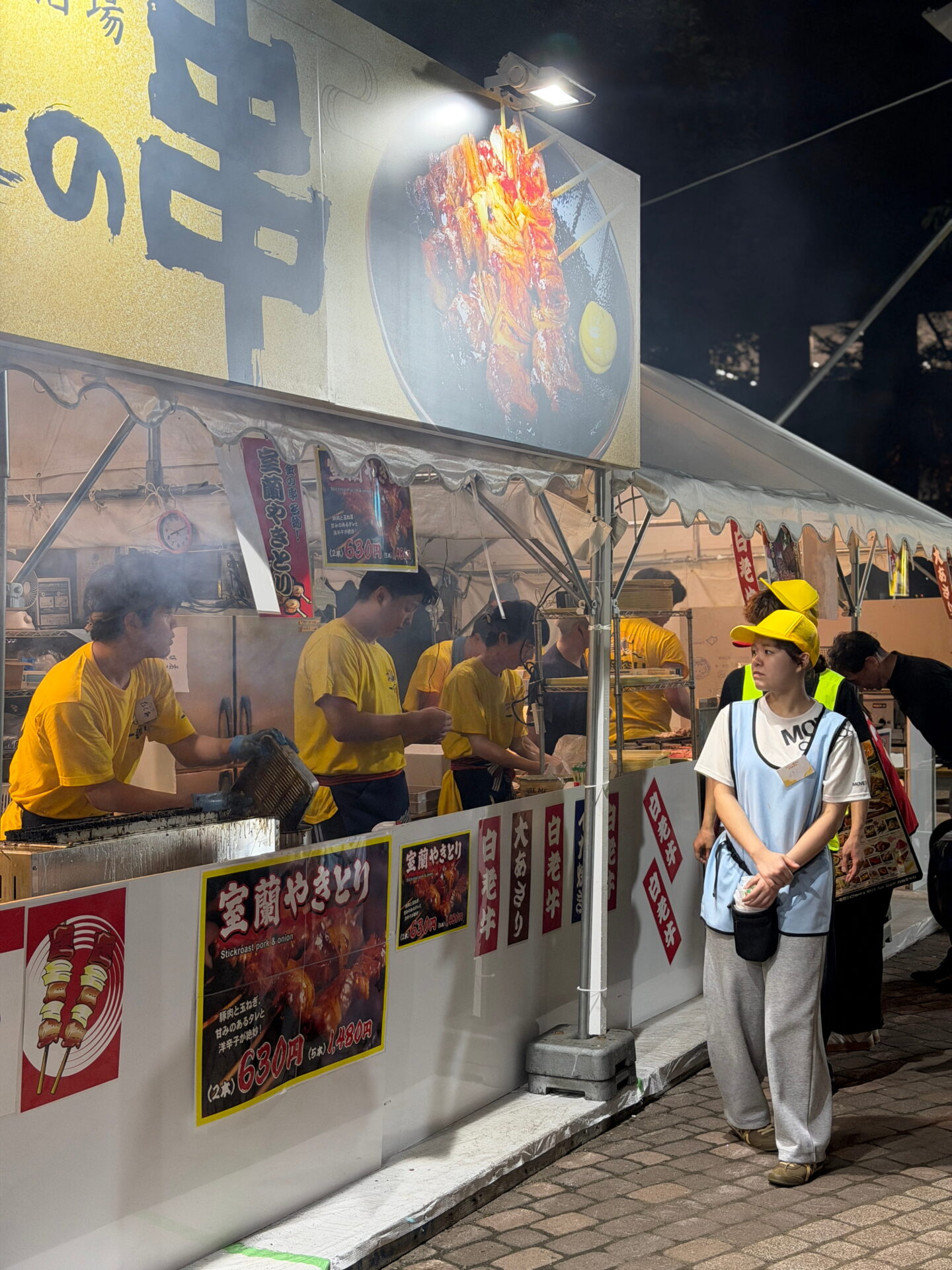 Food vendors in yellow shirts prepare grilled meat skewers at a brightly lit outdoor food stall in Sapporo at night. Japanese signs advertise Muroran yakitori made with pork and onion, with prices starting at 630 yen for two skewers.