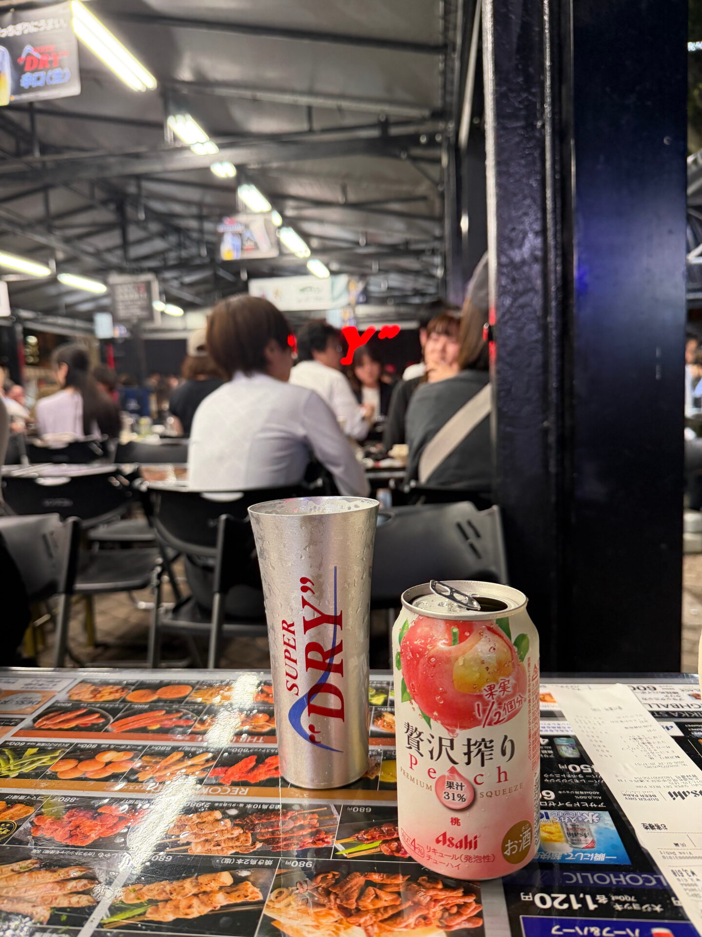 A tall silver Asahi Super Dry beer cup and a pink Asahi peach flavoured alcoholic drink sit on a table covered with Japanese food menus. Diners fill the busy outdoor beer garden in Sapporo beneath a large tent illuminated by overhead lights.