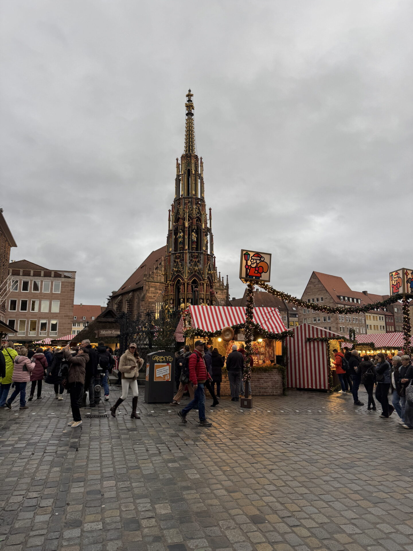 The ornate Gothic Schöner Brunnen fountain rises above Nuremberg Christmas market beneath a grey winter sky. Visitors browse wooden stalls with red and white striped roofs, surrounded by historic buildings and festive garlands.