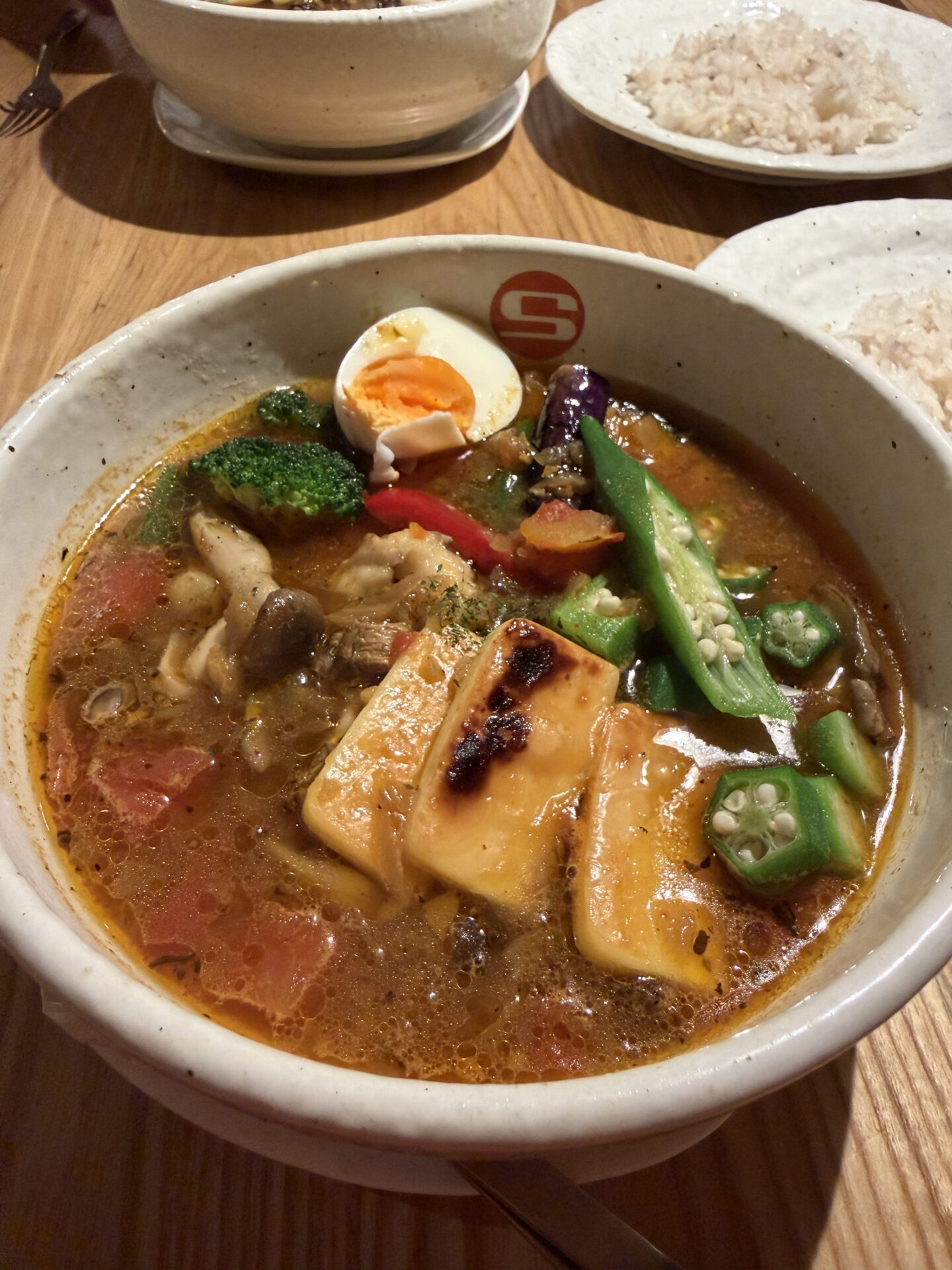 Close up of a steaming bowl of Japanese soup curry in a rich reddish brown broth, topped with grilled cheese, sliced okra, broccoli, mushrooms, red pepper and half a boiled egg. A plate of rice sits behind the bowl on a wooden restaurant table.