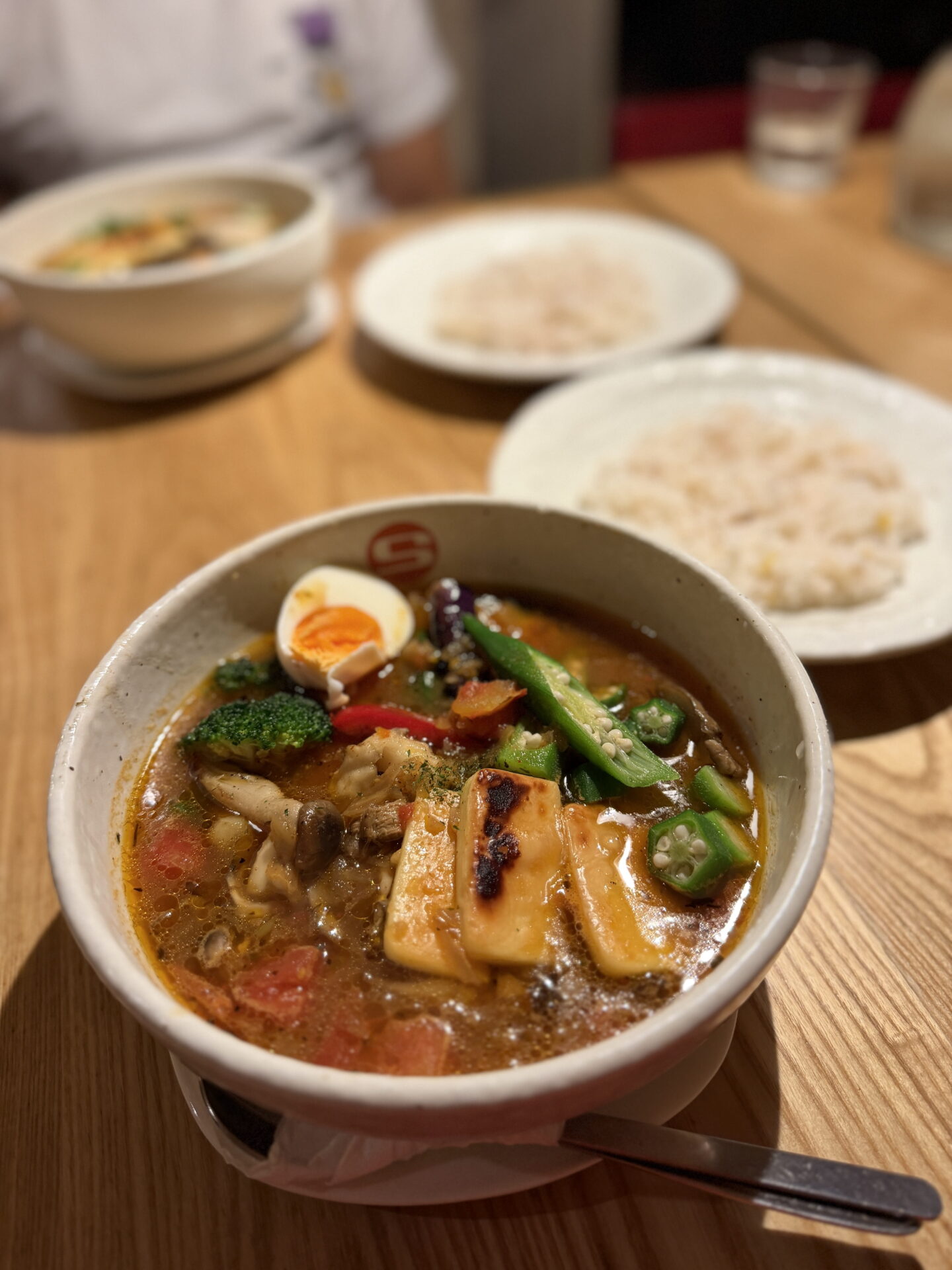 A bowl of Japanese soup curry filled with grilled cheese, green okra, broccoli, mushrooms, tomatoes and half a boiled egg in a spiced broth. Two plates of rice and another bowl of soup curry sit blurred in the background.